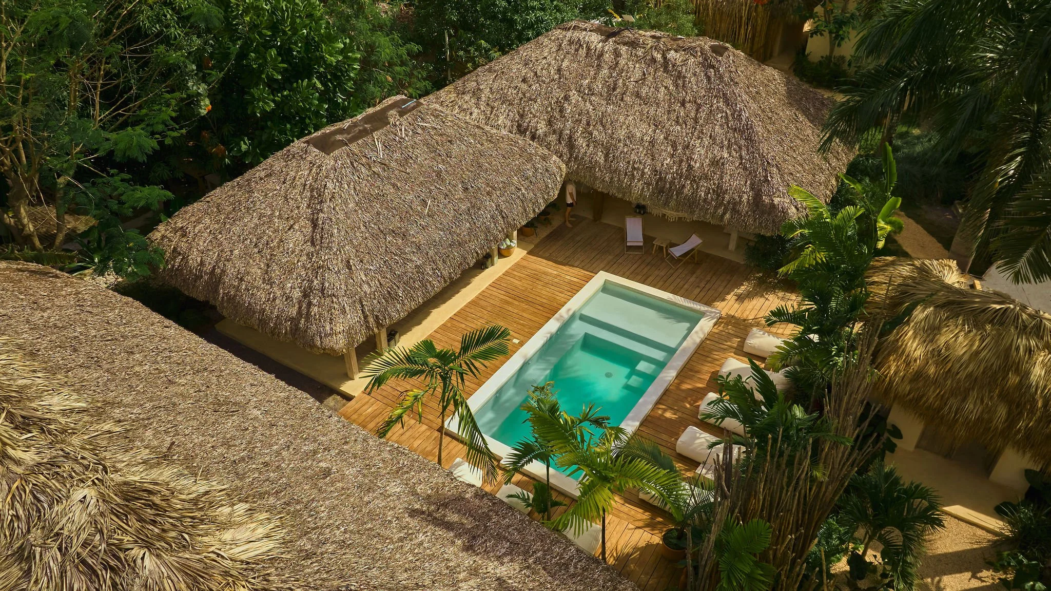 Vista aérea de un área de piscina rodeada de estructura de techo de palma y vegetación tropical, con área de descanso y sombra en un ambiente de playa o resort tropical.