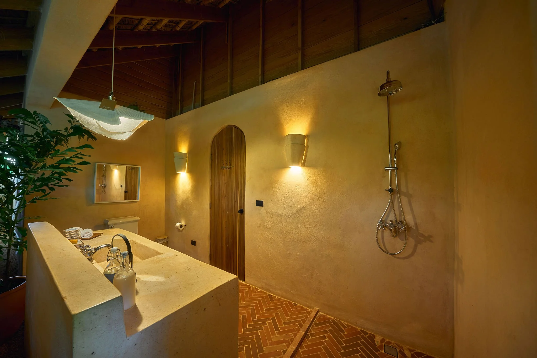 Baño con paredes de color arena, lavabo de piedra, espejo, luces empotradas y ducha con cabeza de lluvia, decorado con plantas.