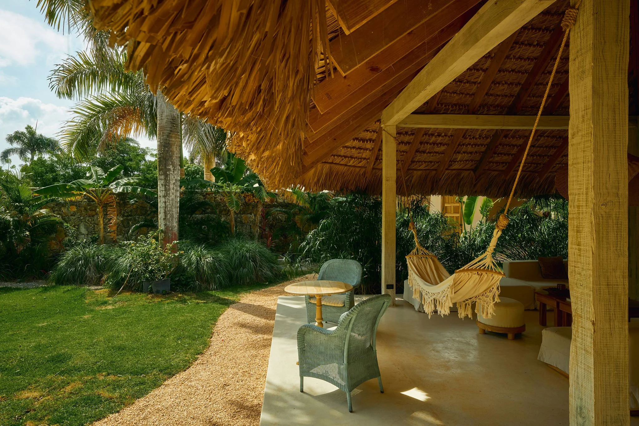Porche cubierto con mobiliario de jardín y hamaca, rodeado de vegetación tropical y palmeras bajo cielo parcialmente nublado.