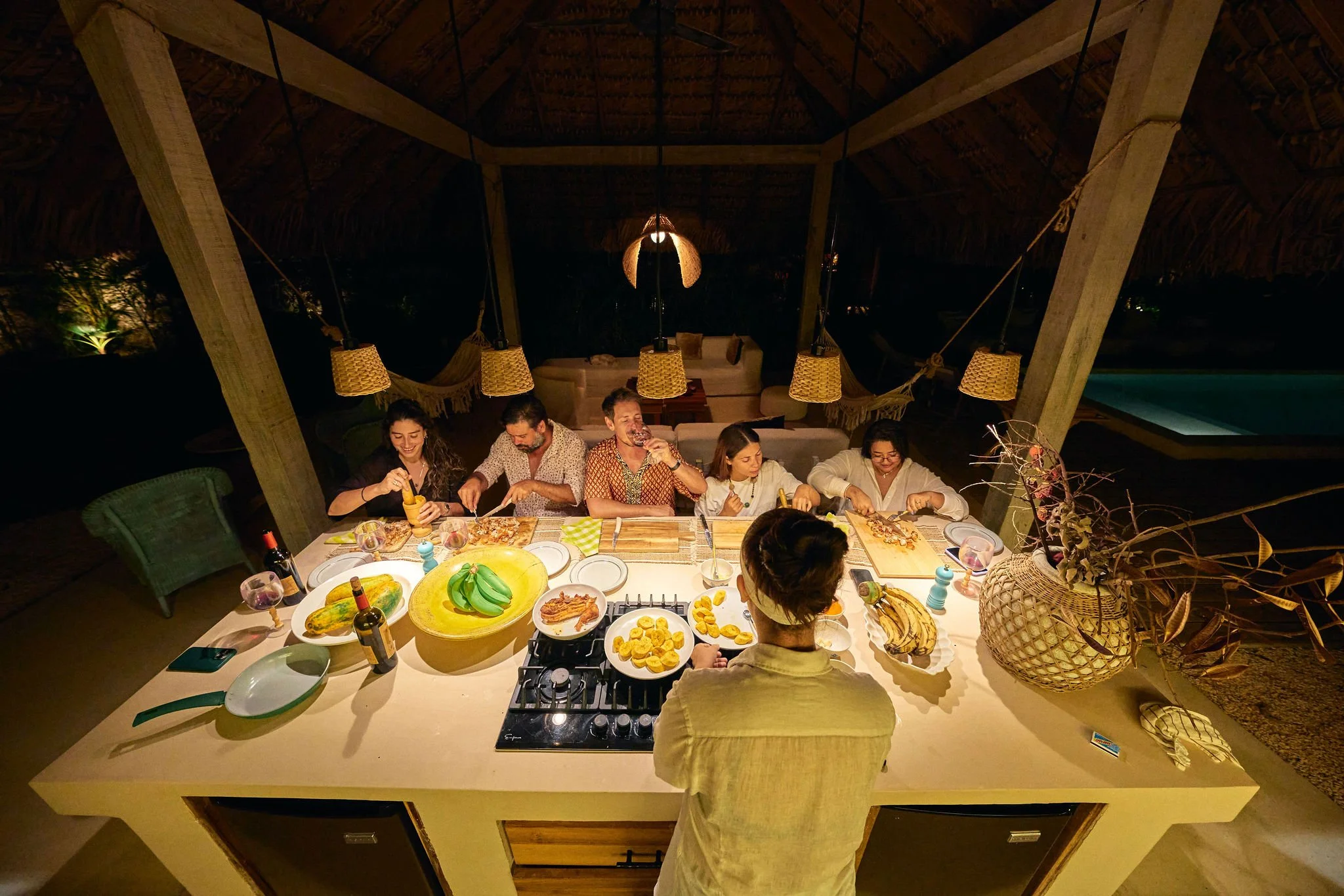 Grupo de personas cenando en una mesa al aire libre en la noche, con comida y utensilios sobre la mesa, decorada con lámparas, plantas y un ambiente acogedor.