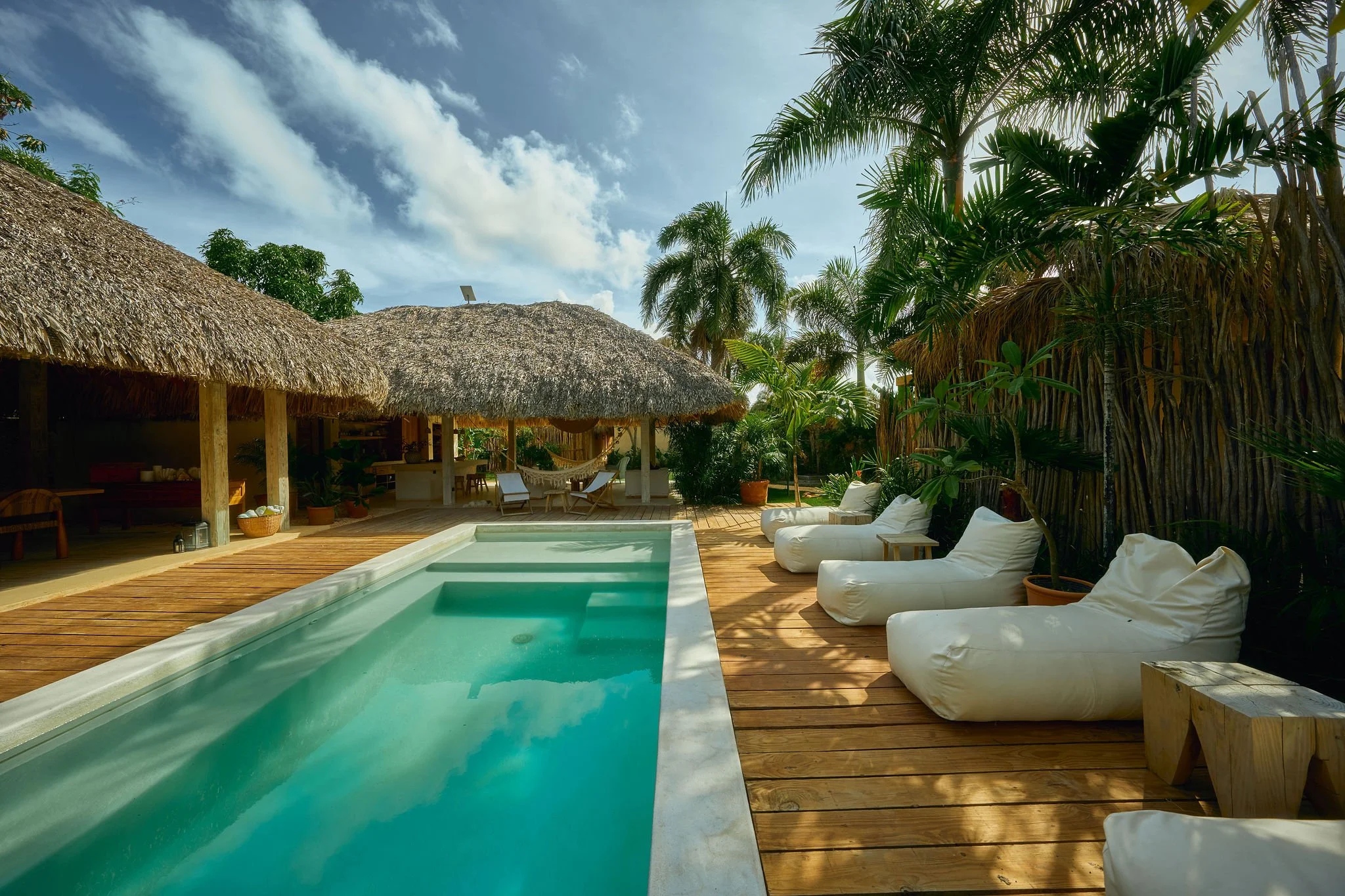 Piscina en un área de descanso con sillas blancas y palmeras, casas con techos de paja y cielo con nubes.
