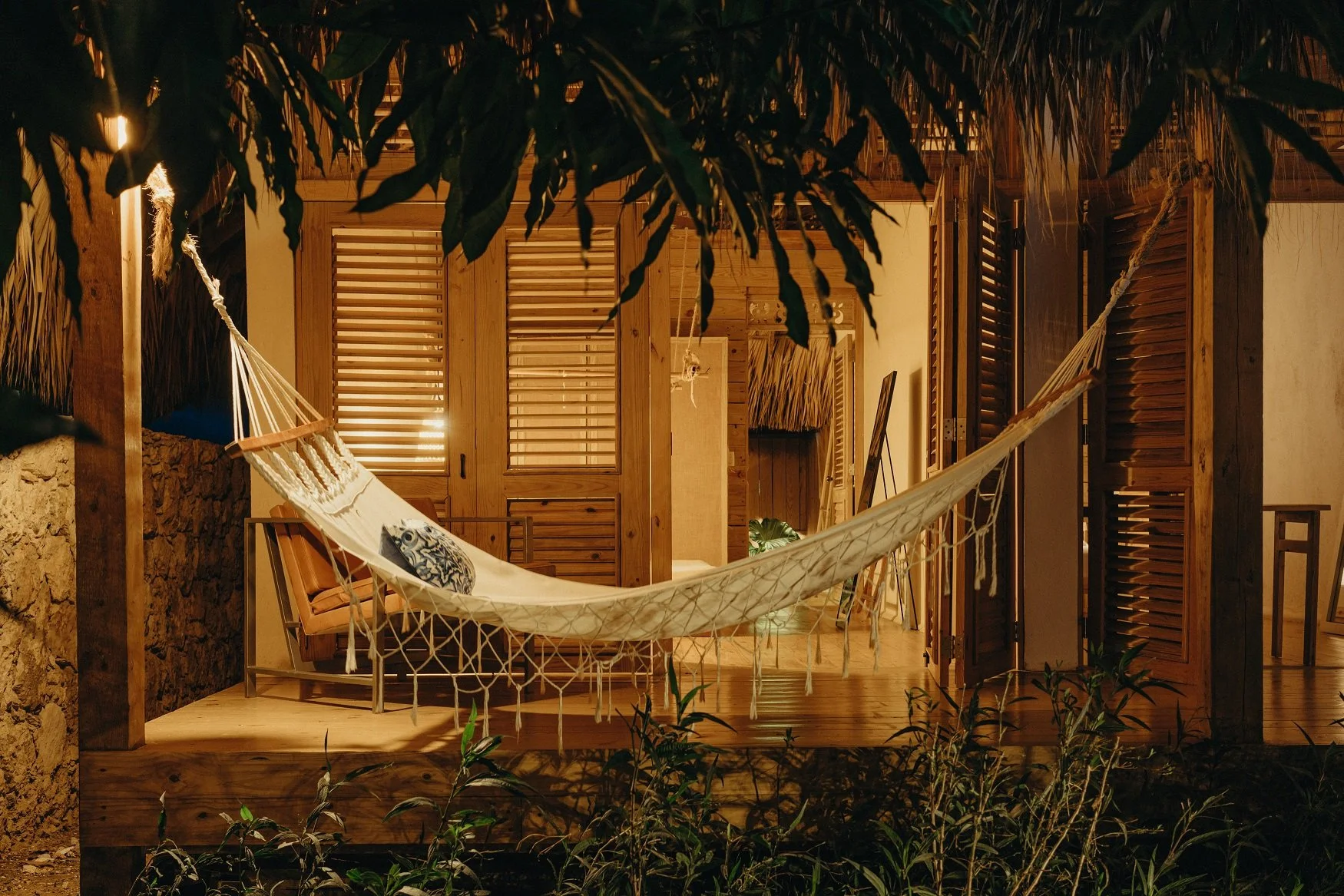 Terraza de madera con una hamaca, cojines y plantas decorativas, iluminada cálidamente por la noche.