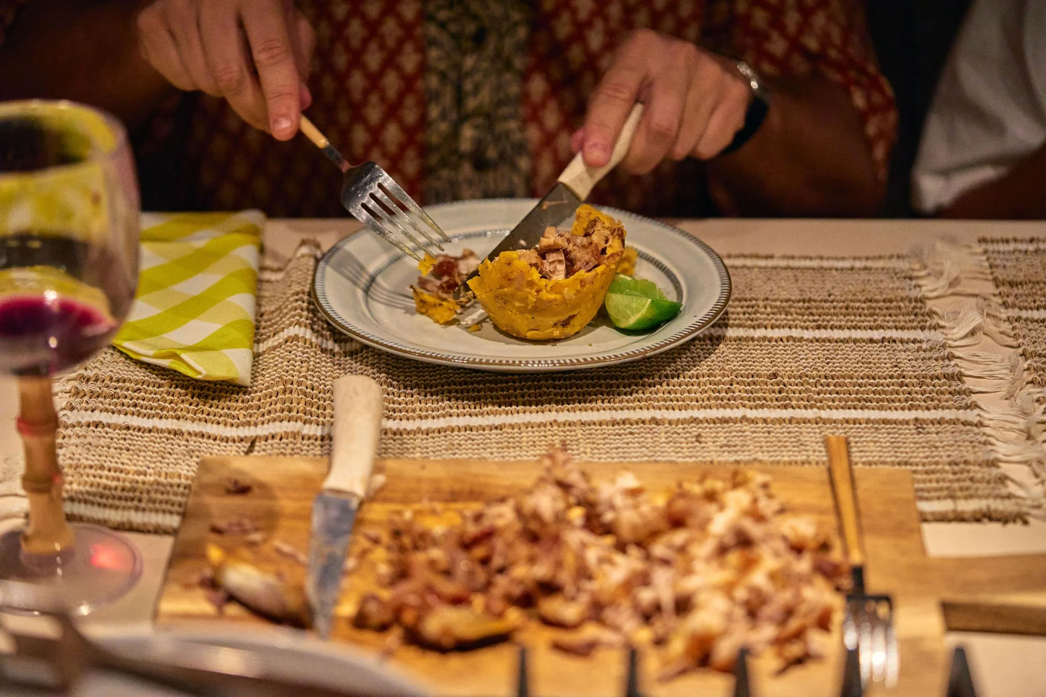 Persona sirviendo comida en un plato con lima, en una mesa decorada con mantel de yute y platito. En primer plano, carne asada en un plato de madera y vino tinto.