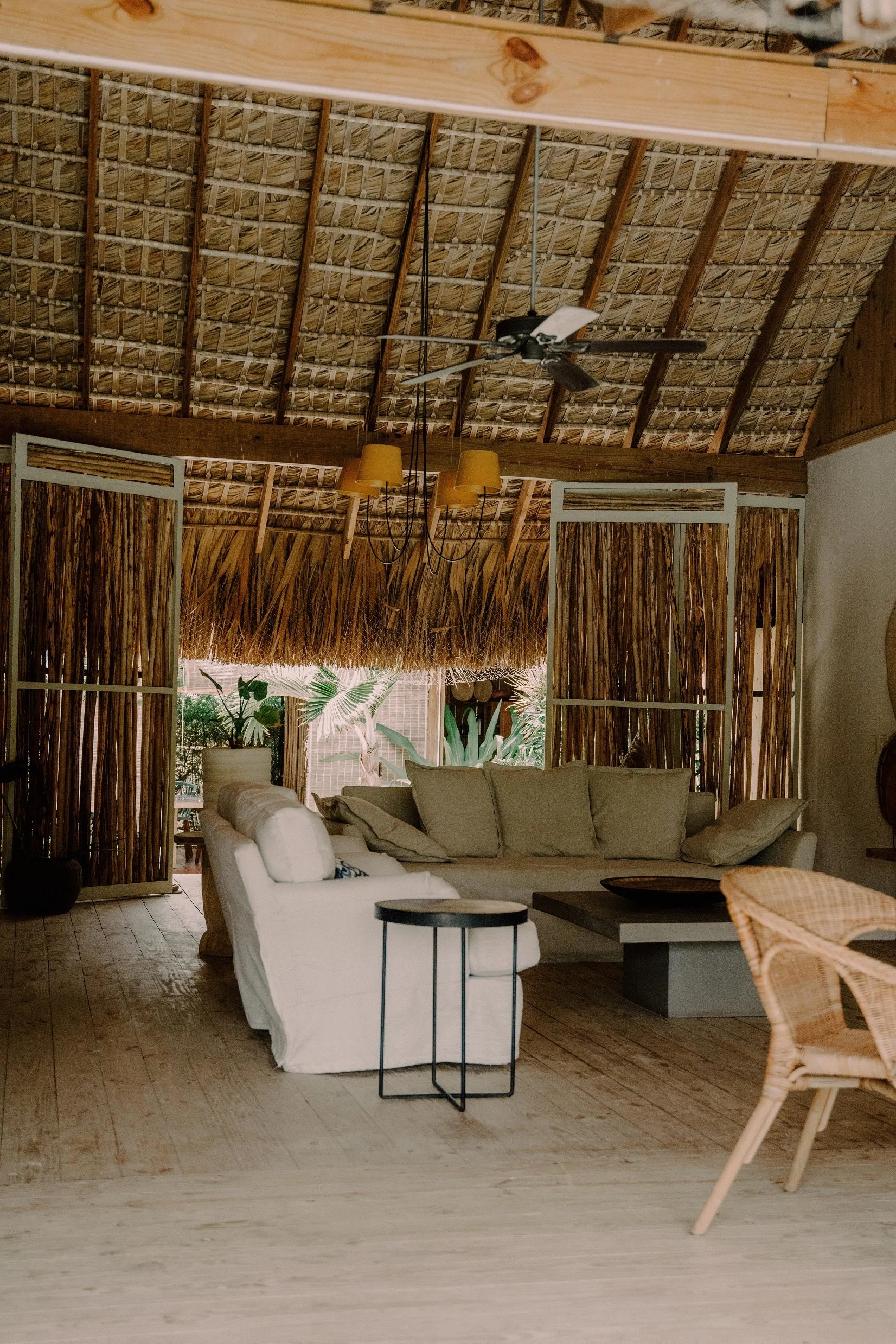 Sala decorada con muebles en un ambiente rústico con puertas de madera de caña, sofás beige, una mesa de centro de concreto, una pequeña mesa negra, sillones de mimbre, plantas tropicales y techo de palapa.