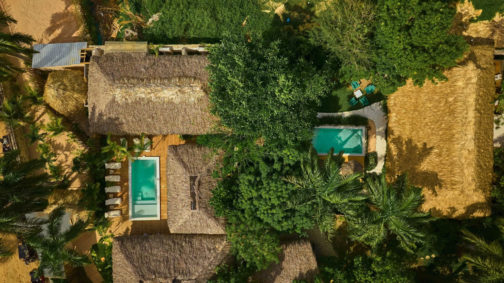 Vista aérea de una propiedad con varias cabañas de techo de paja, dos piscinas pequeñas, árboles verdes y mobiliario de exterior, en un entorno tropical.