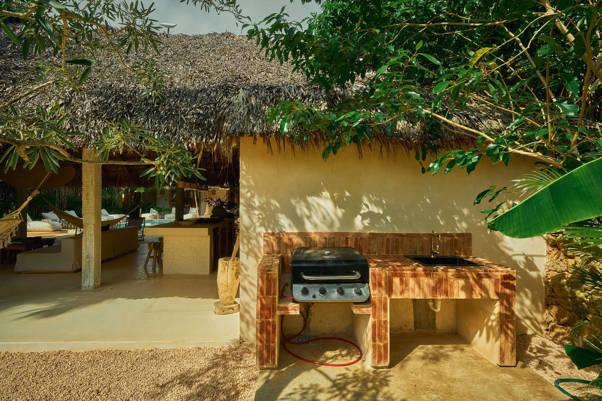Área al aire libre con cocina de piedra, parrilla y fregadero, sombra de árboles y plantas verdes, estructura de esteras de palma en fondo, mobiliario de descanso y piscina en el fondo.