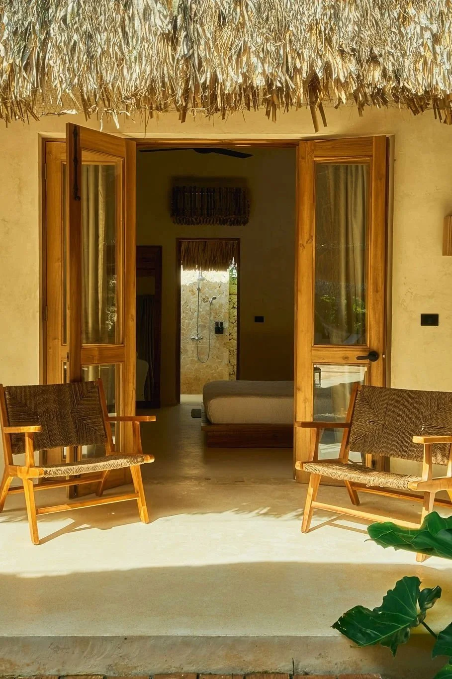 Vista frontal de una casa de playa con puertas abiertas de madera, dos sillas de madera con asientos de cuerda y plantas en primer plano.