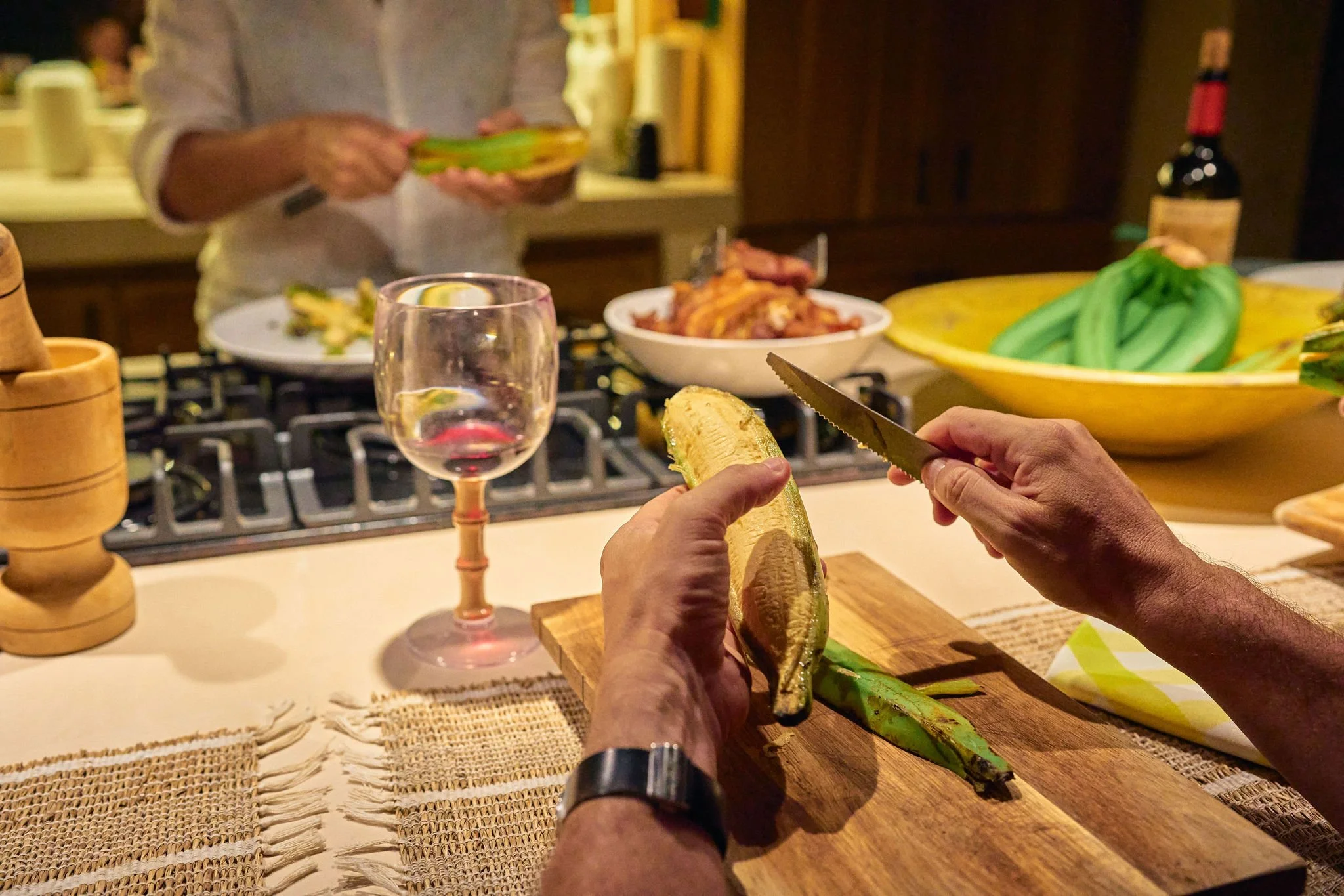 Personas pelando plátanos en una cocina, con comida y vino en la mesa.