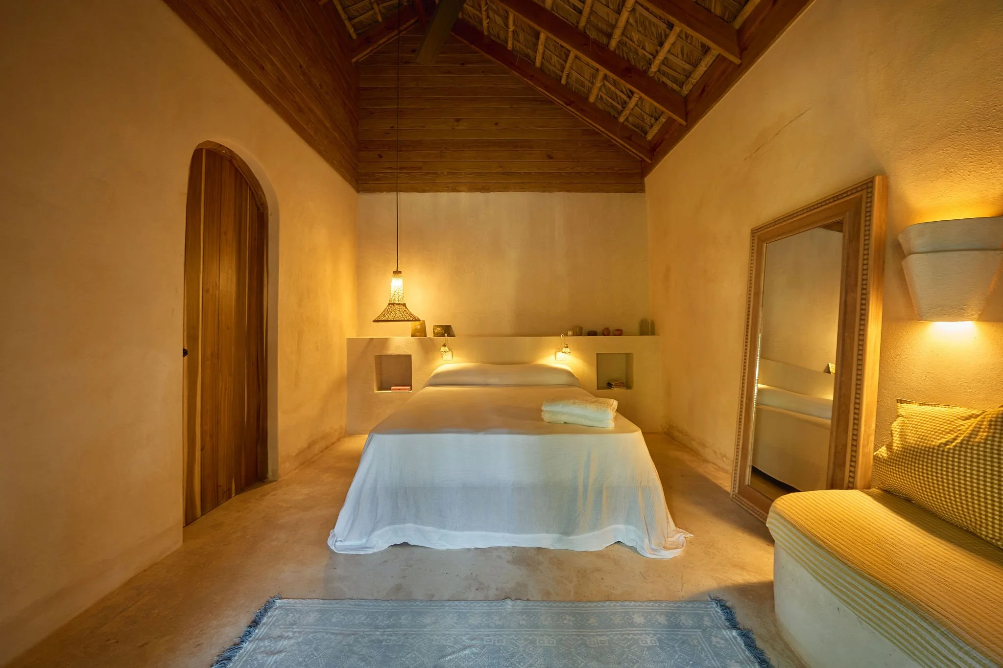 Habitación con techo de madera, cama blanca, espejo de piso y lámparas de pared en tonos cálidos.