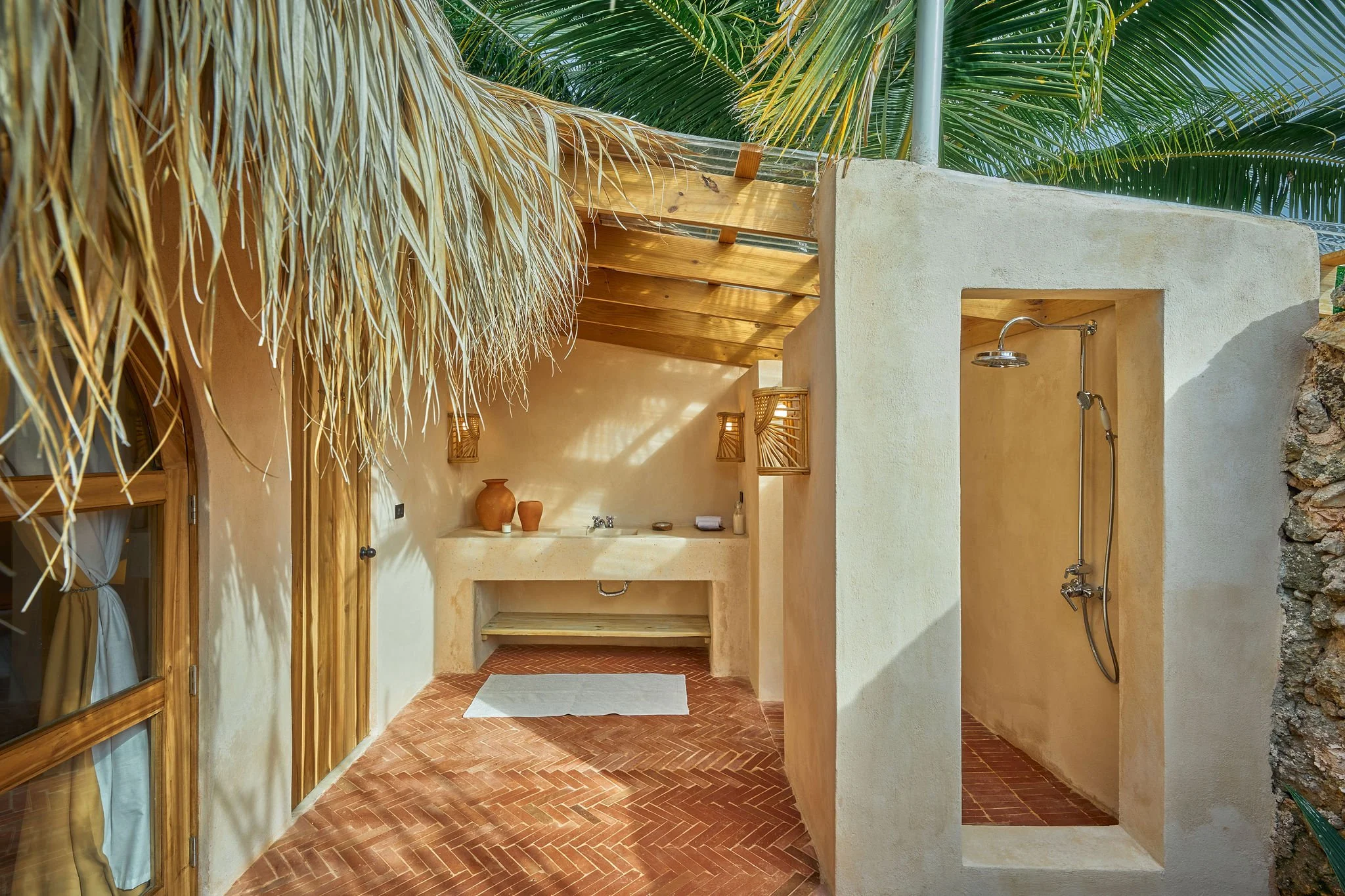 Ducha exterior con paredes de yeso, ducha de lluvia, techo de madera, decoraciones sencillas con vasijas de barro, suelo de ladrillo y toldo de palma