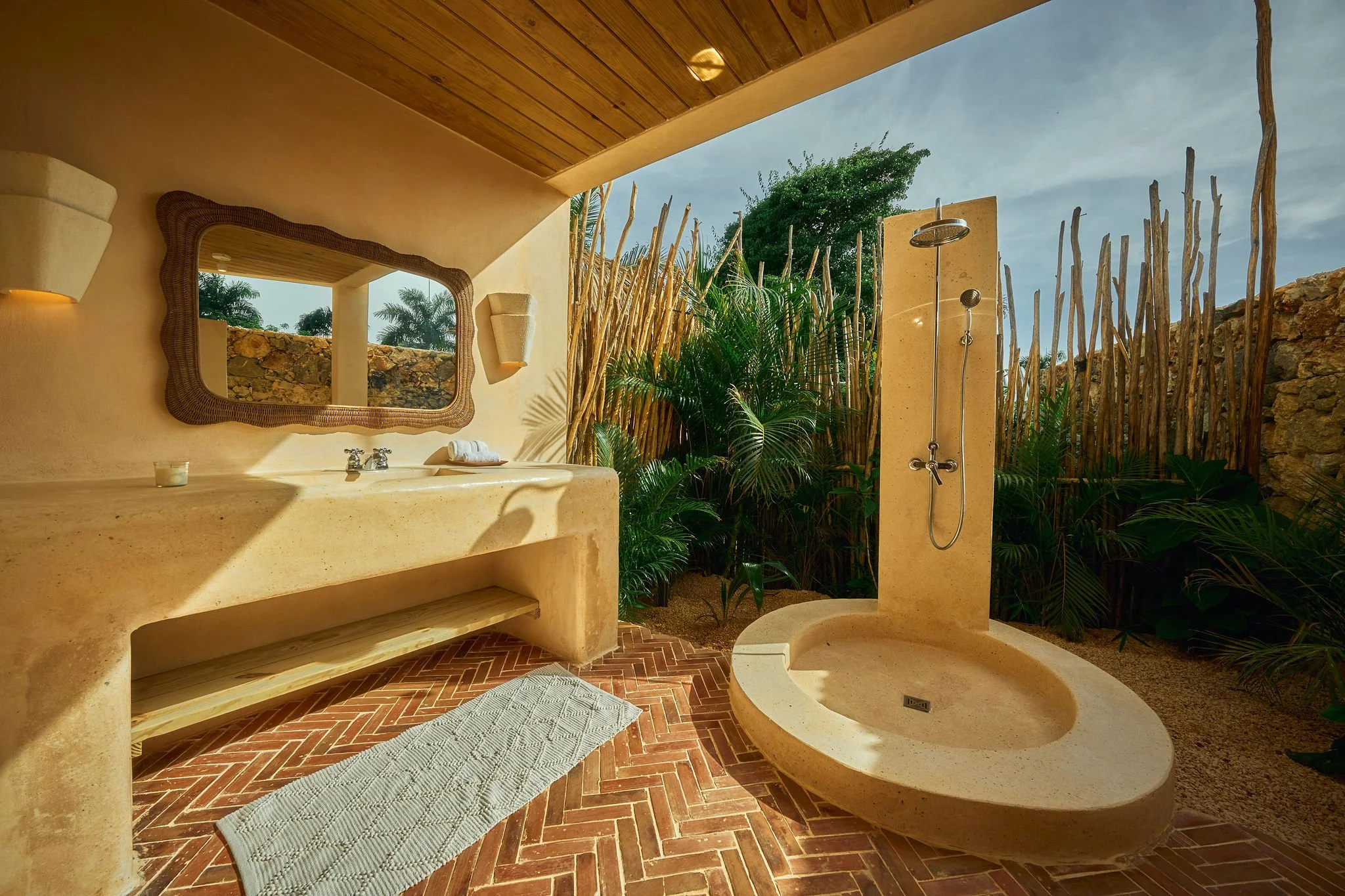 Bañera al aire libre y ducha en un baño con paredes de piedra y plantas tropicales, ambiente natural y soleado.