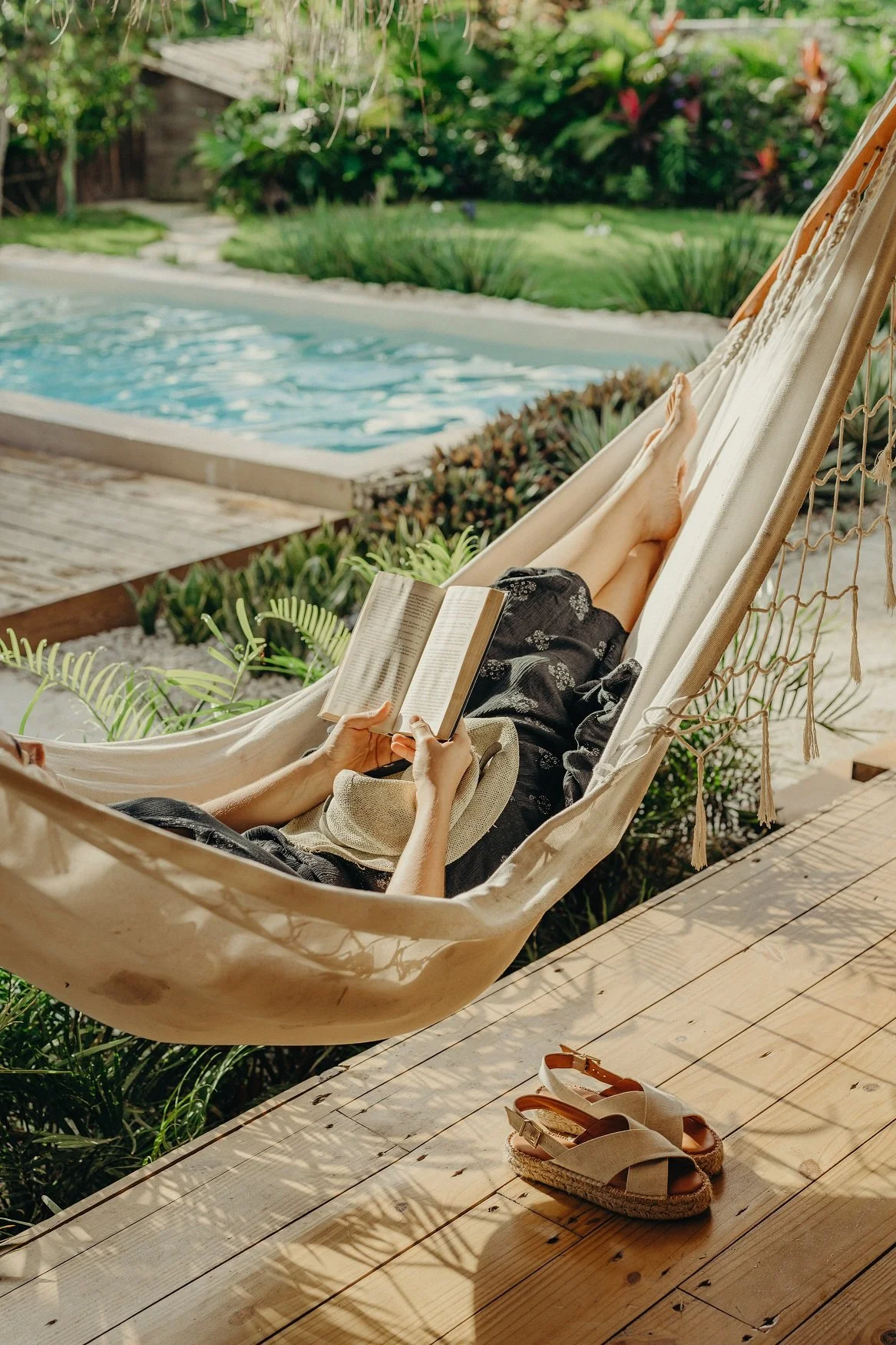 Persona descansando en una hamaca leyendo un libro cerca de una piscina en un jardín con plantas y sombra, con sandalias de yute en el suelo de madera.
