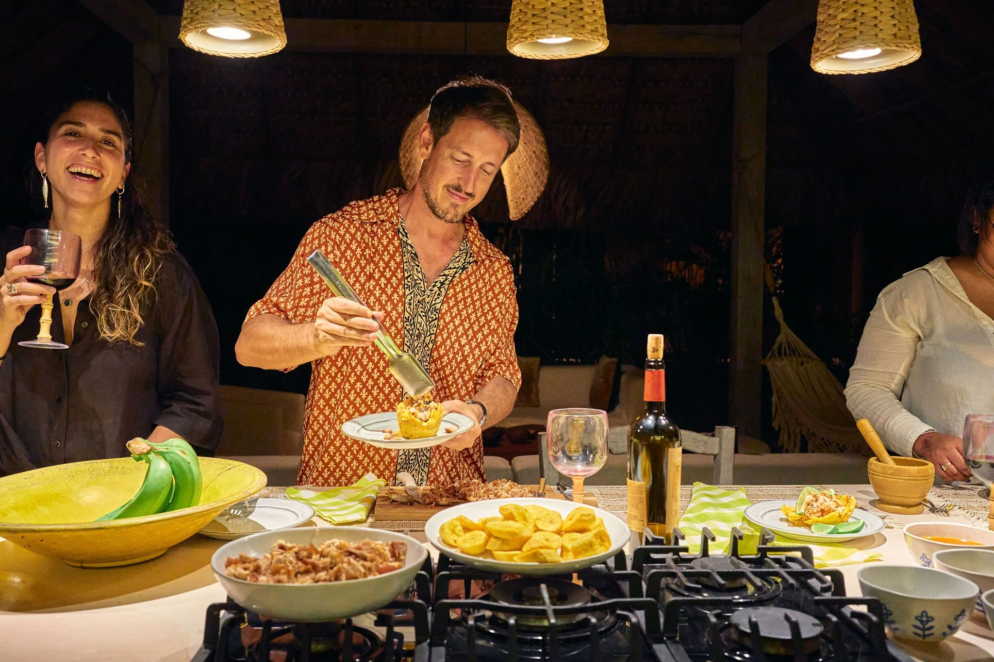 Personas en una cena preparando comida en una mesa con platos, vino y comida, en un ambiente acogedor.