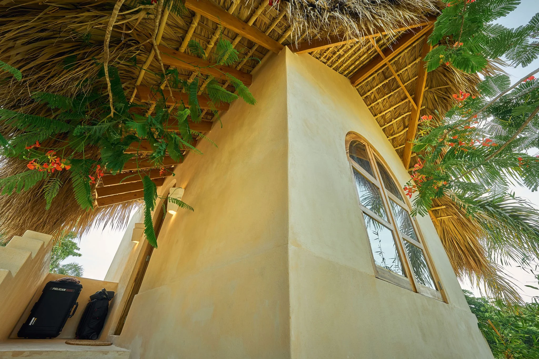 Vista desde abajo de una casa de estilo maya con paredes amarillas, ventanas de madera, techo de palma y plantas con flores naranjas alrededor.