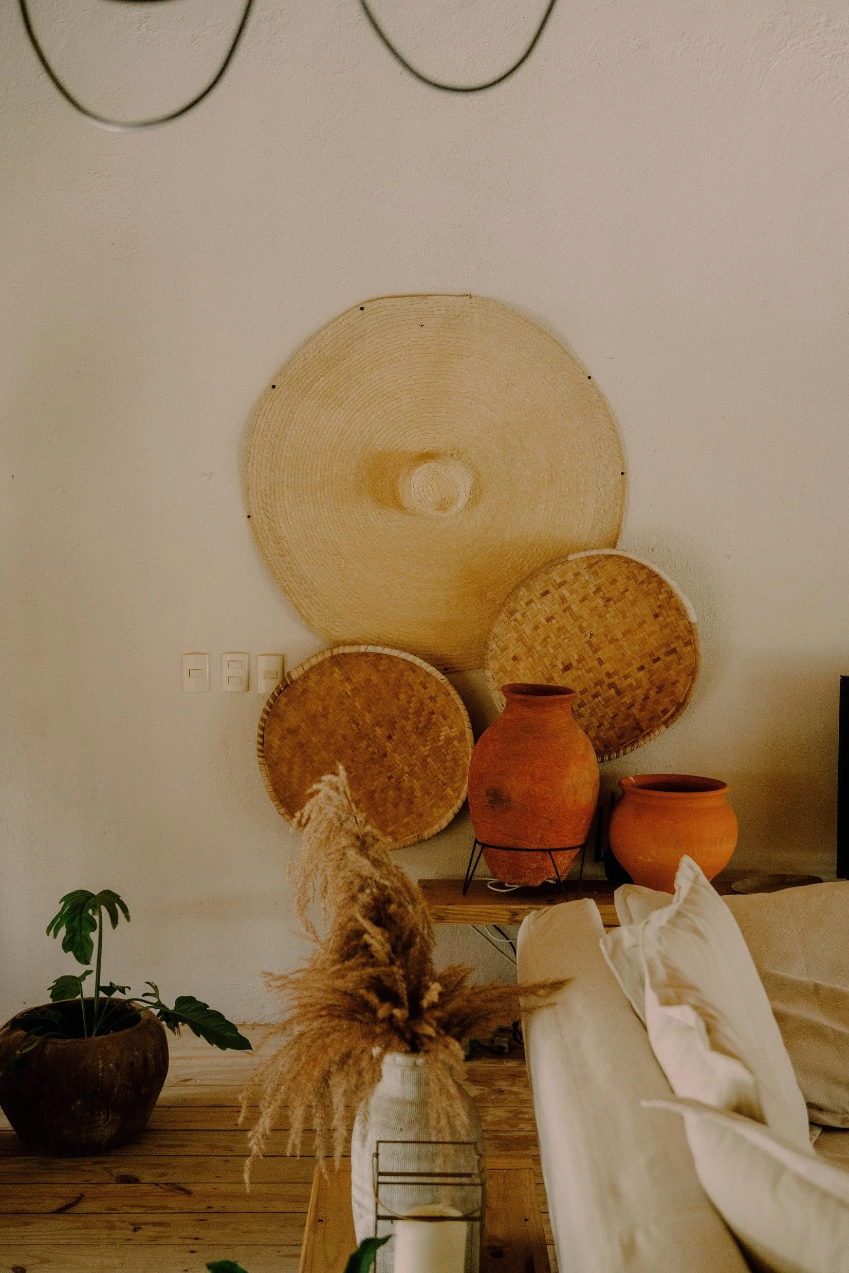 Decoración de interior con cestas de mimbre de diferentes tamaños en la pared, vasijas de barro en la mesa, plantas en macetas y un sofá de color claro.