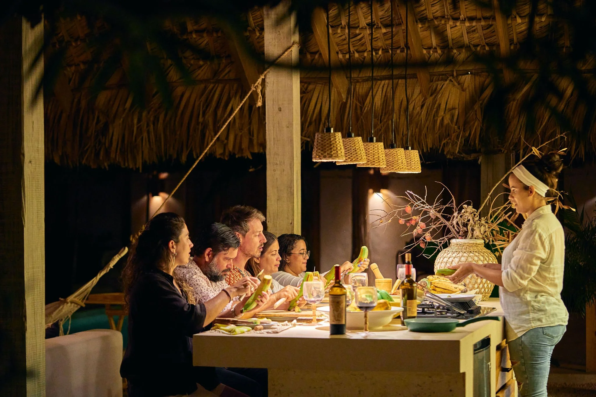 Grupo de personas en una comida al aire libre en un espacio decorado con estructuras de techo de hoja de palma, siendo atendidas por una camarera que presenta alimentos.