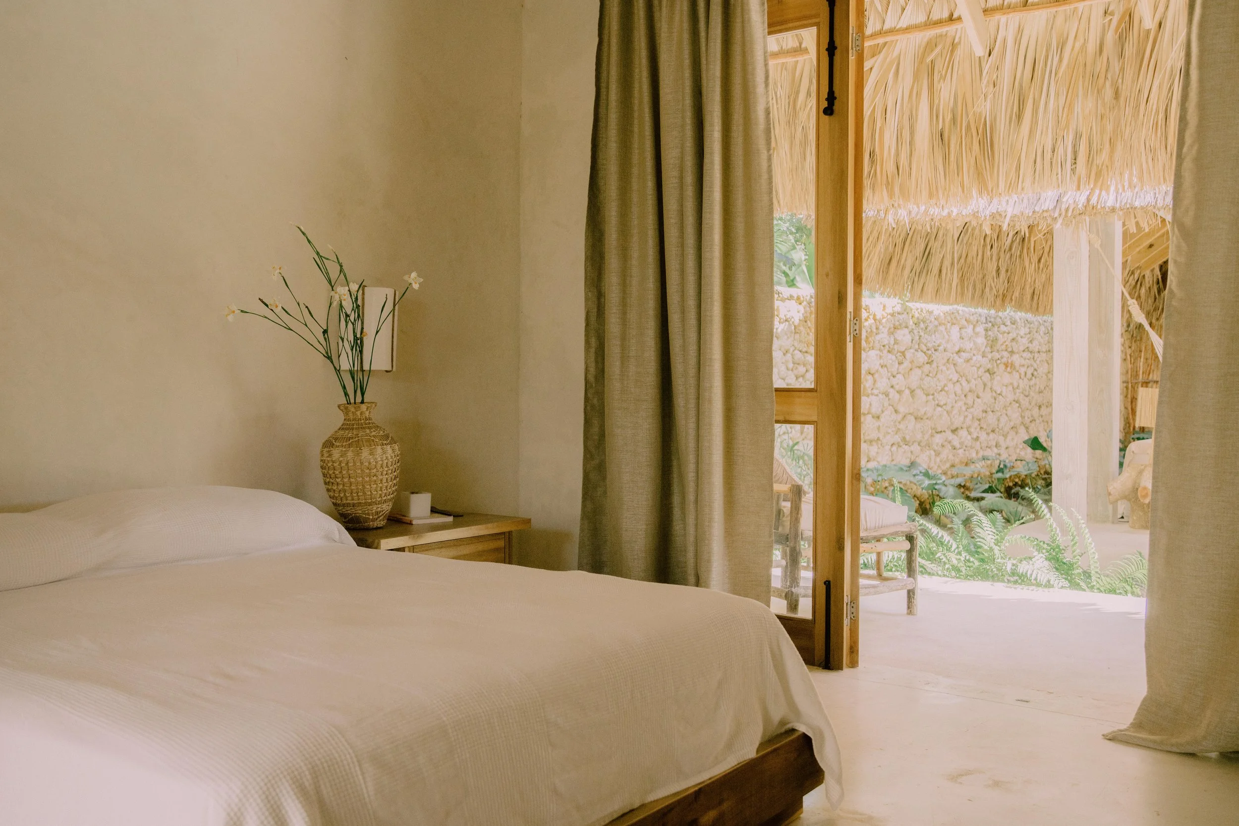 Habitación con cama blanca, mesita de noche, jarrón de mimbre con flores y vista a un espacio exterior con paredes de piedra y techo de palma.