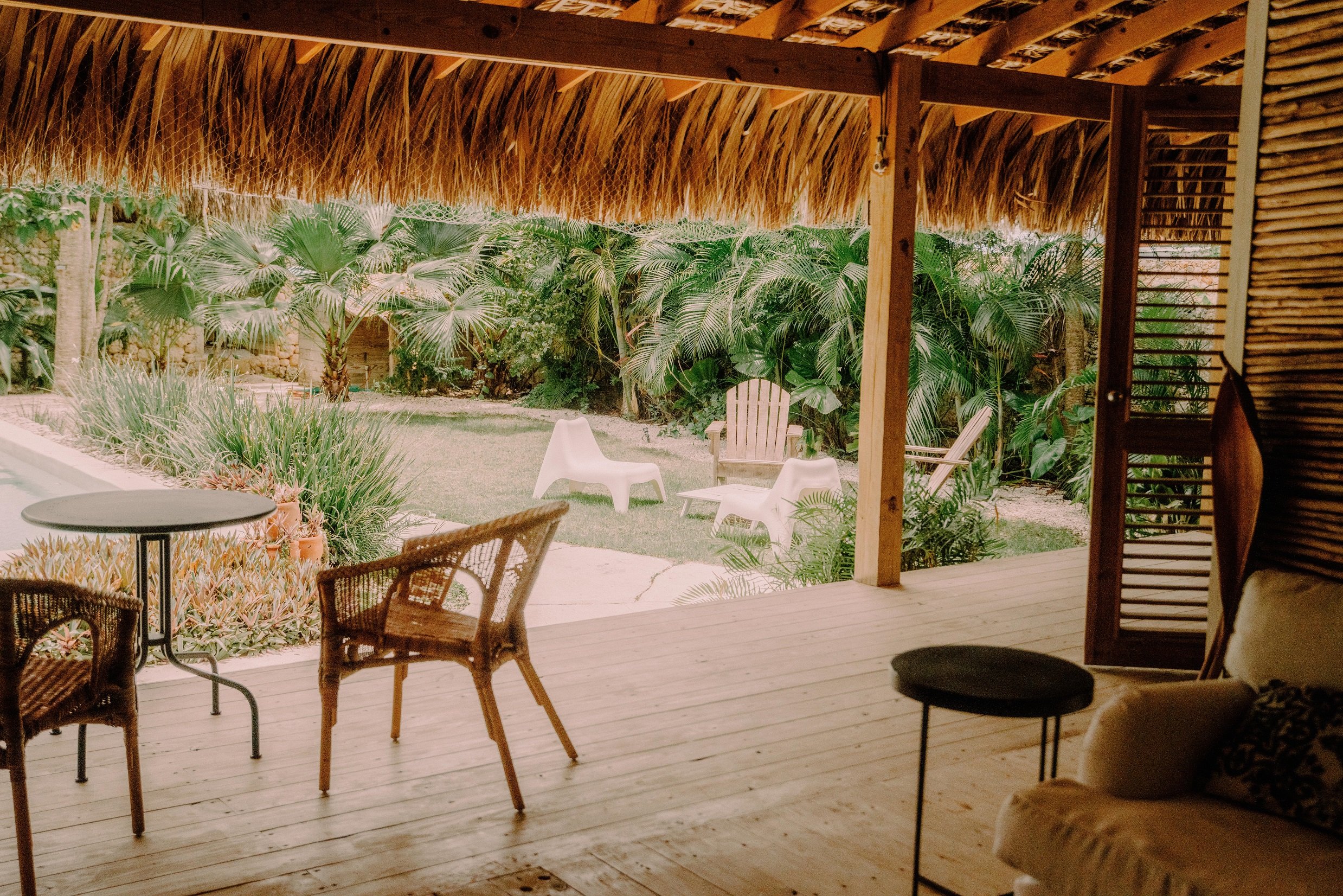 Vista desde una sala con piso de madera, sillón, mesa pequeña y sillas de mimbre, hacia un jardín con plantas tropicales y sillas blancas.
