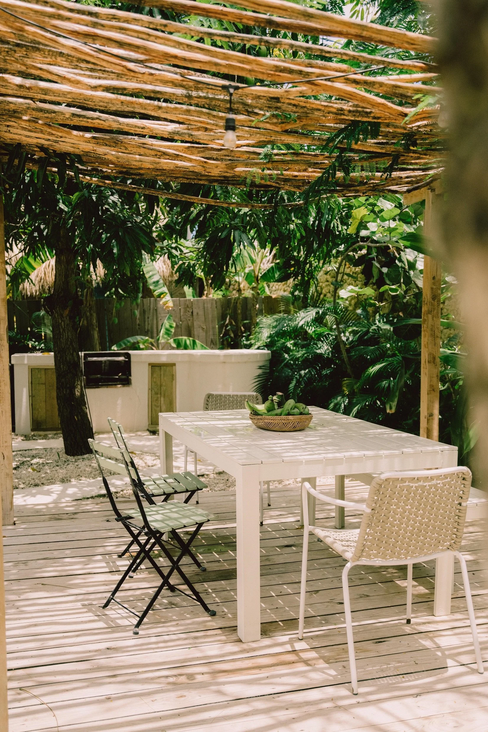 Un área de comedor al aire libre con mesa blanca y sillas, rodeada de vegetación y sombra, bajo un techo de cañas de bambú.