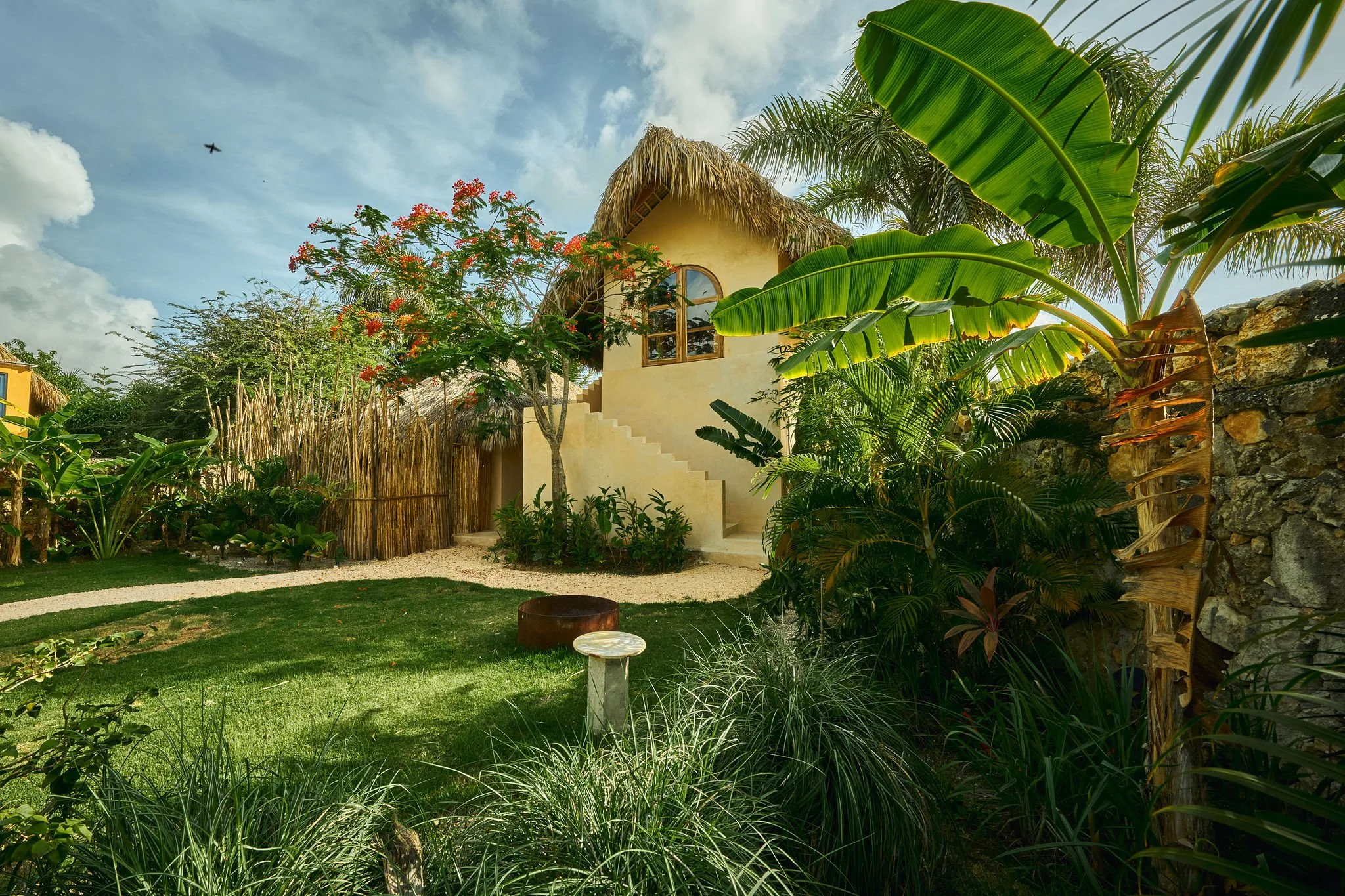 Casa de estilo rústico en un jardín rodeado de plantas tropicales y árboles, con un cielo despejado y un avión en el fondo.