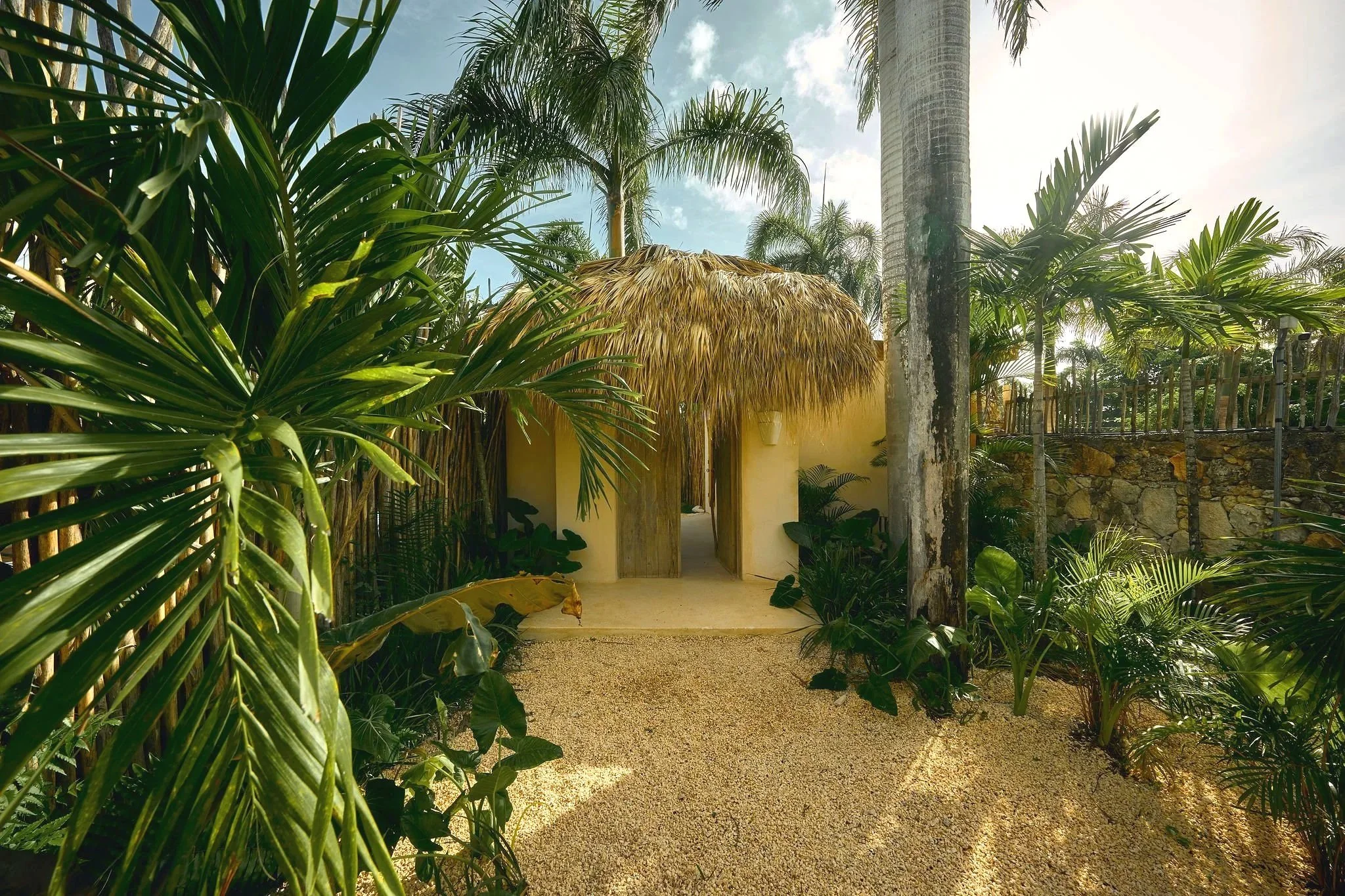 Entrada de una casa con techo de paja, rodeada de plantas tropicales y palmeras, con camino de gravilla y muro de piedra a la derecha