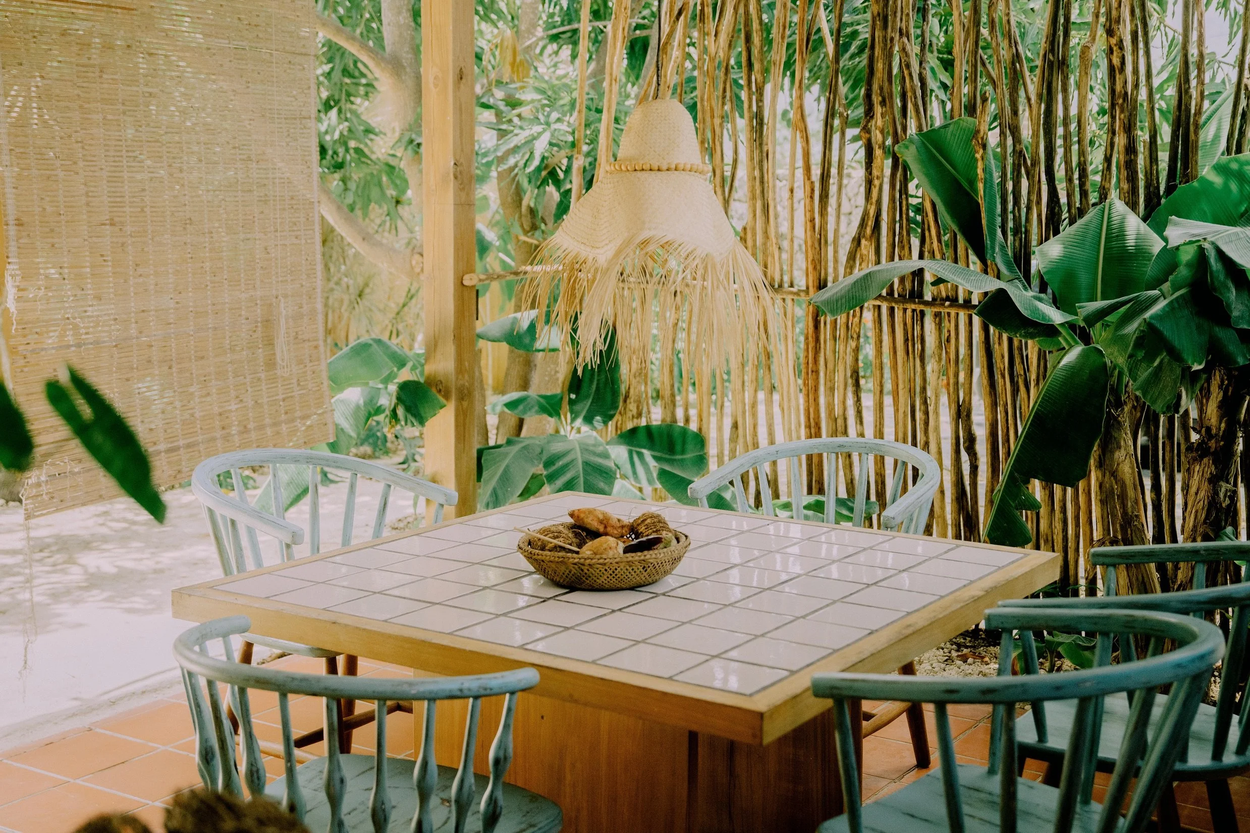 Imagen de un comedor en un espacio al aire libre con sillones blancos y una mesa con azulejos blancos en el centro, decorada con un cesto de pan, rodeado de plantas verdes y una pared de madera.