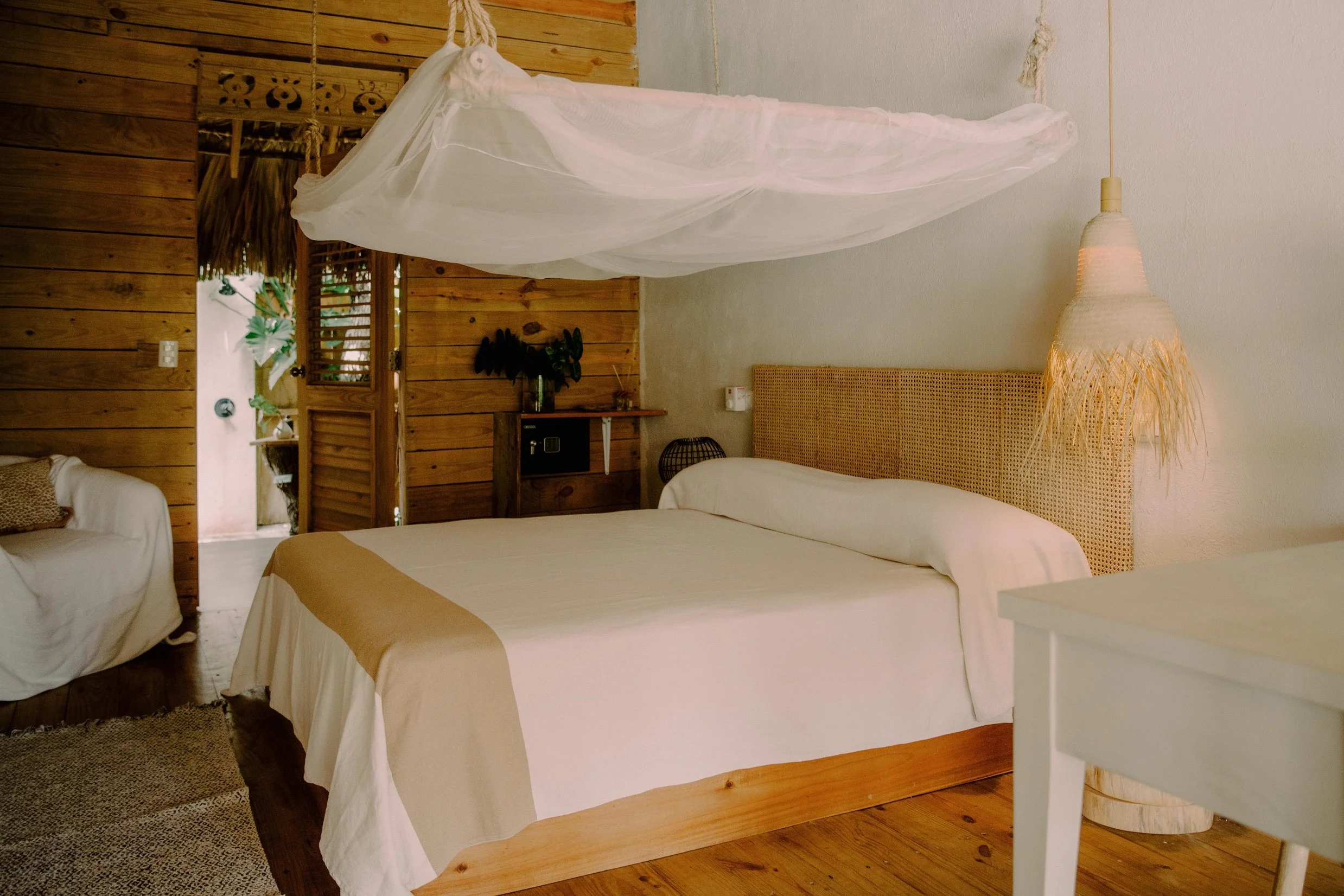 Habitación con cama grande, cabecera de ratán, lámpara de tejido y mosquitero colgado del techo, paredes de madera y una puerta de madera que lleva a un área exterior con plantas.