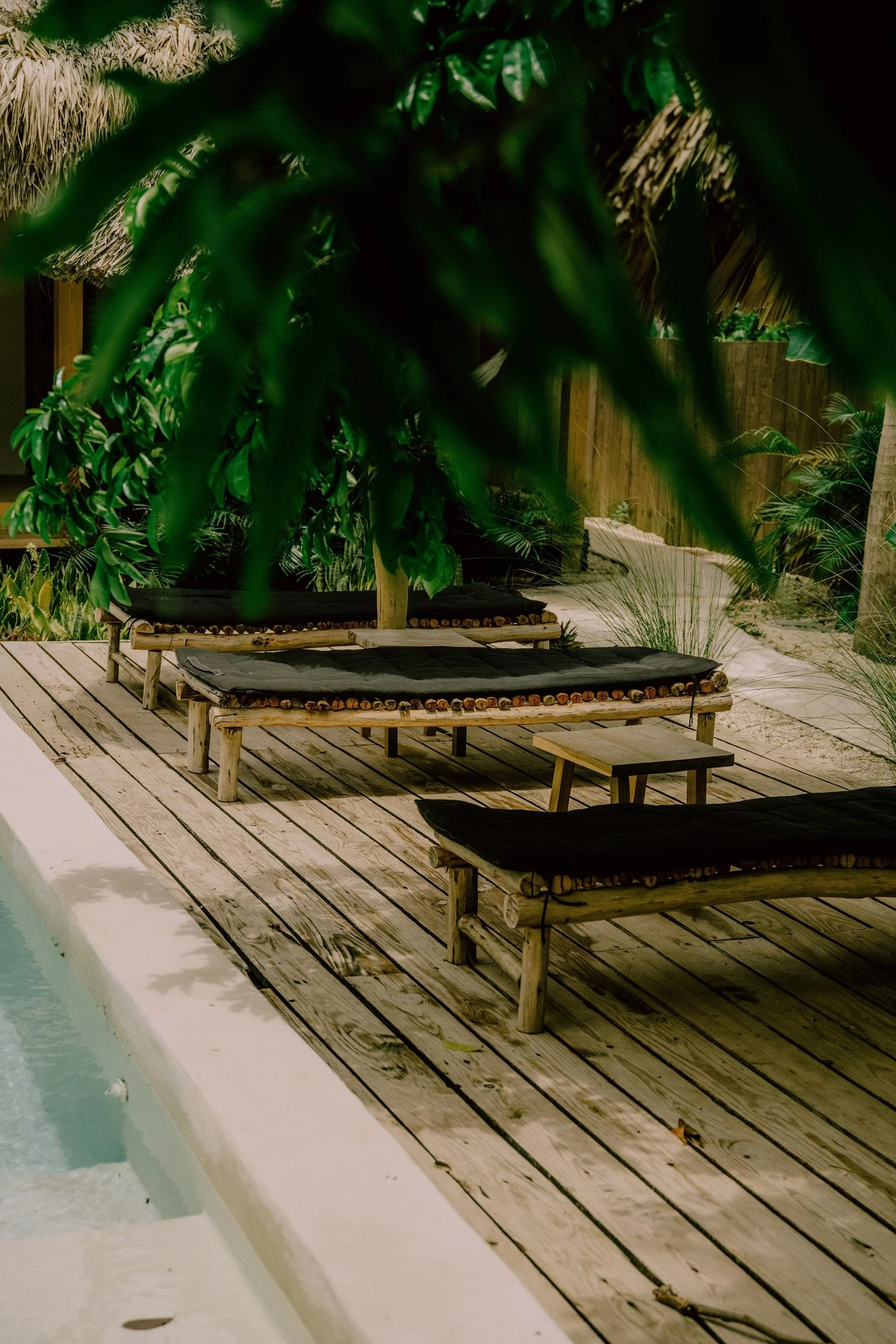 Muebles de madera con cojines negros en una terraza de estilo tropical, rodeada de plantas verdes y una cerca de madera.