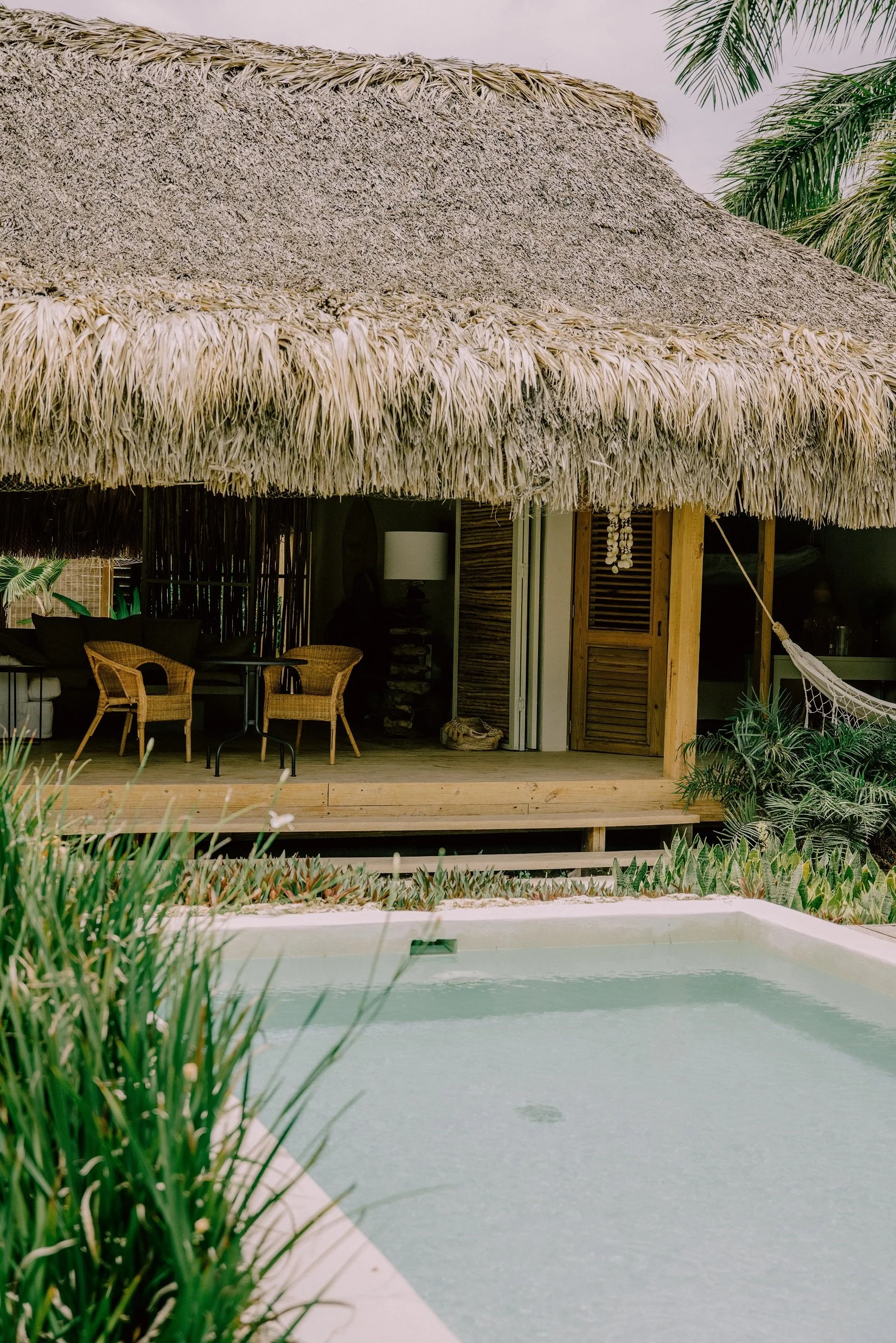 Casa de playa con techo de paja, terraza de madera y piscina cerca de árboles tropicales.