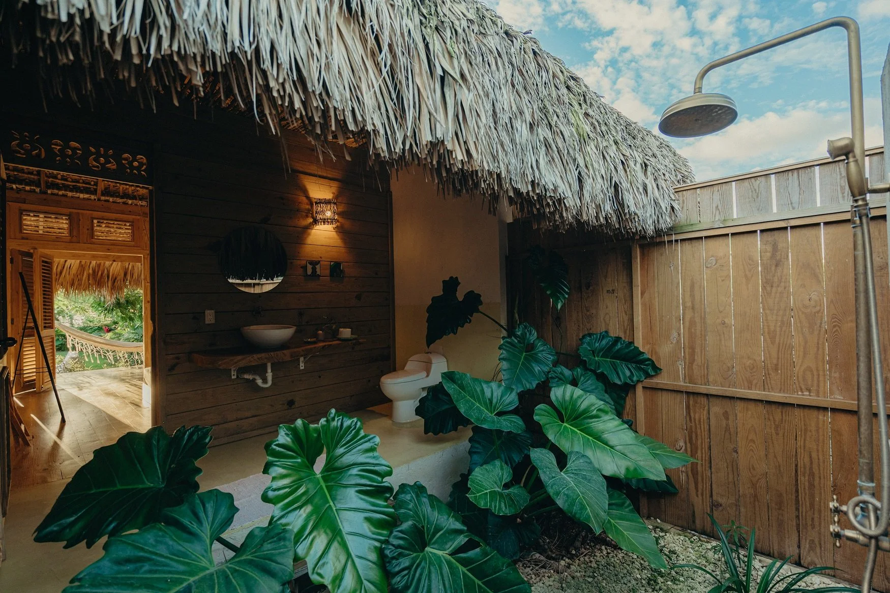 Baño exterior con ducha, lavabo y plantas tropicales, paredes de madera y techo de paja.