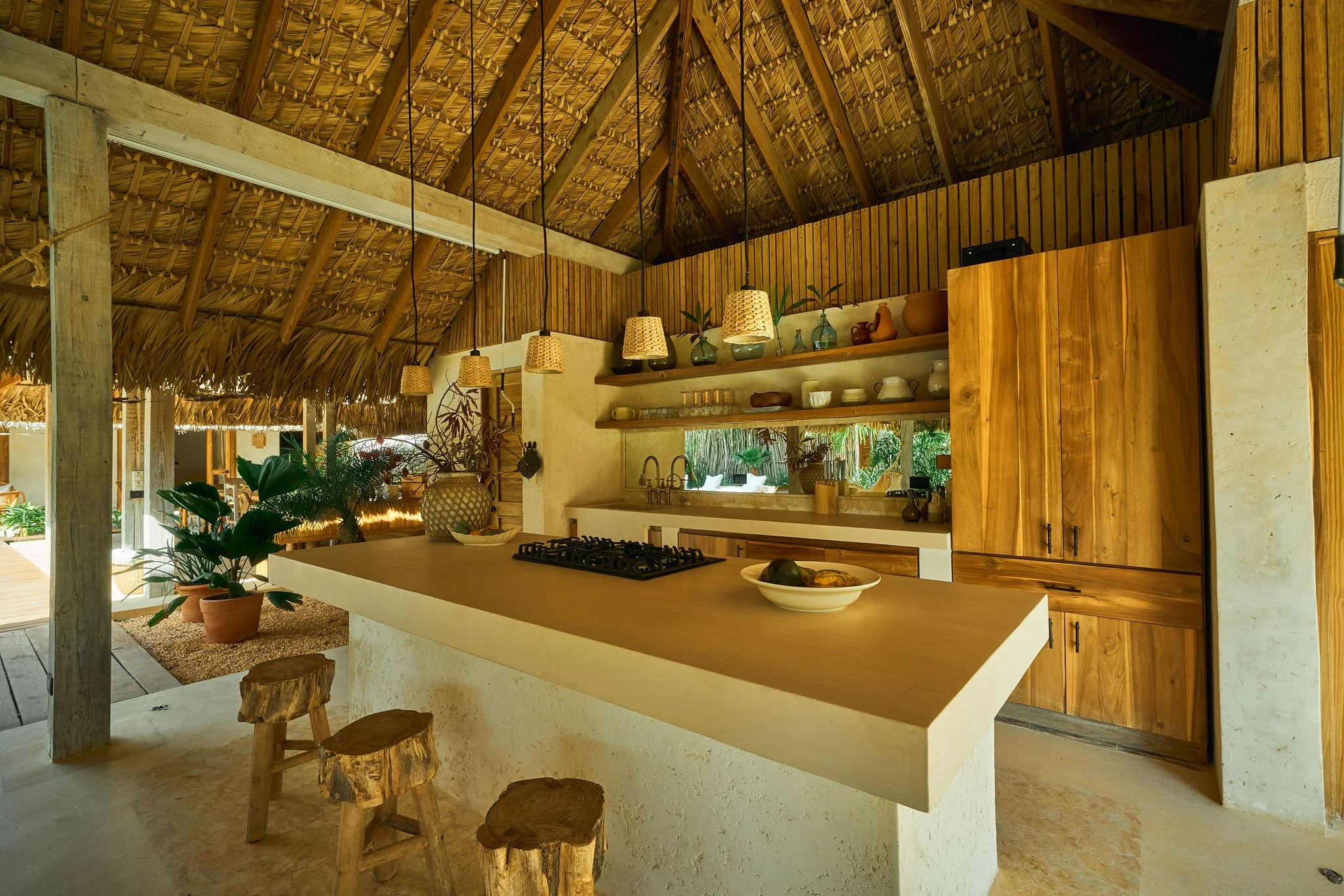 Cocina rústica con techo de paja y paredes de piedra, encimera de concreto, estantes con vajilla y plantas, isla central de madera con utensilios, iluminación de lámparas de mimbre, adornos de cerámica y madera.