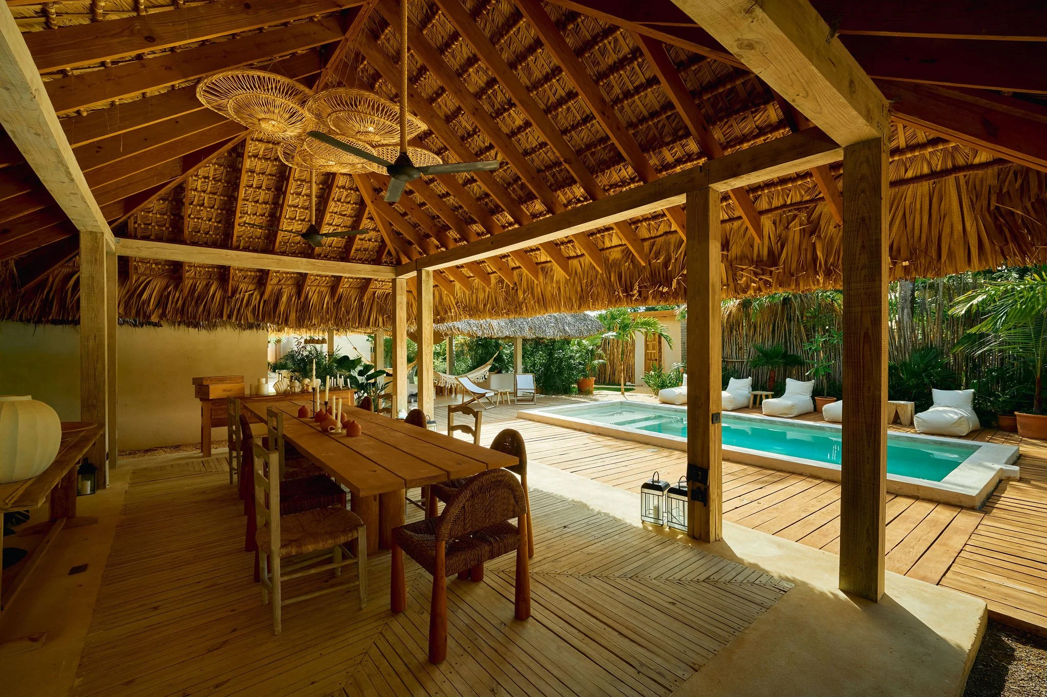 Interior de una casa de playa con estructura de madera, área de comedor, piscina y zona de descanso con hamacas y sillones rodeados de plantas tropicales.