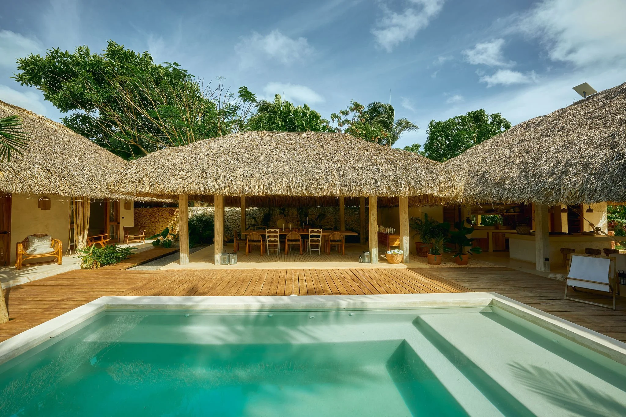 Área de descanso con cabañas de techo de paja, piscina y mobiliario de madera, rodeado de naturaleza tropical bajo cielo despejado.
