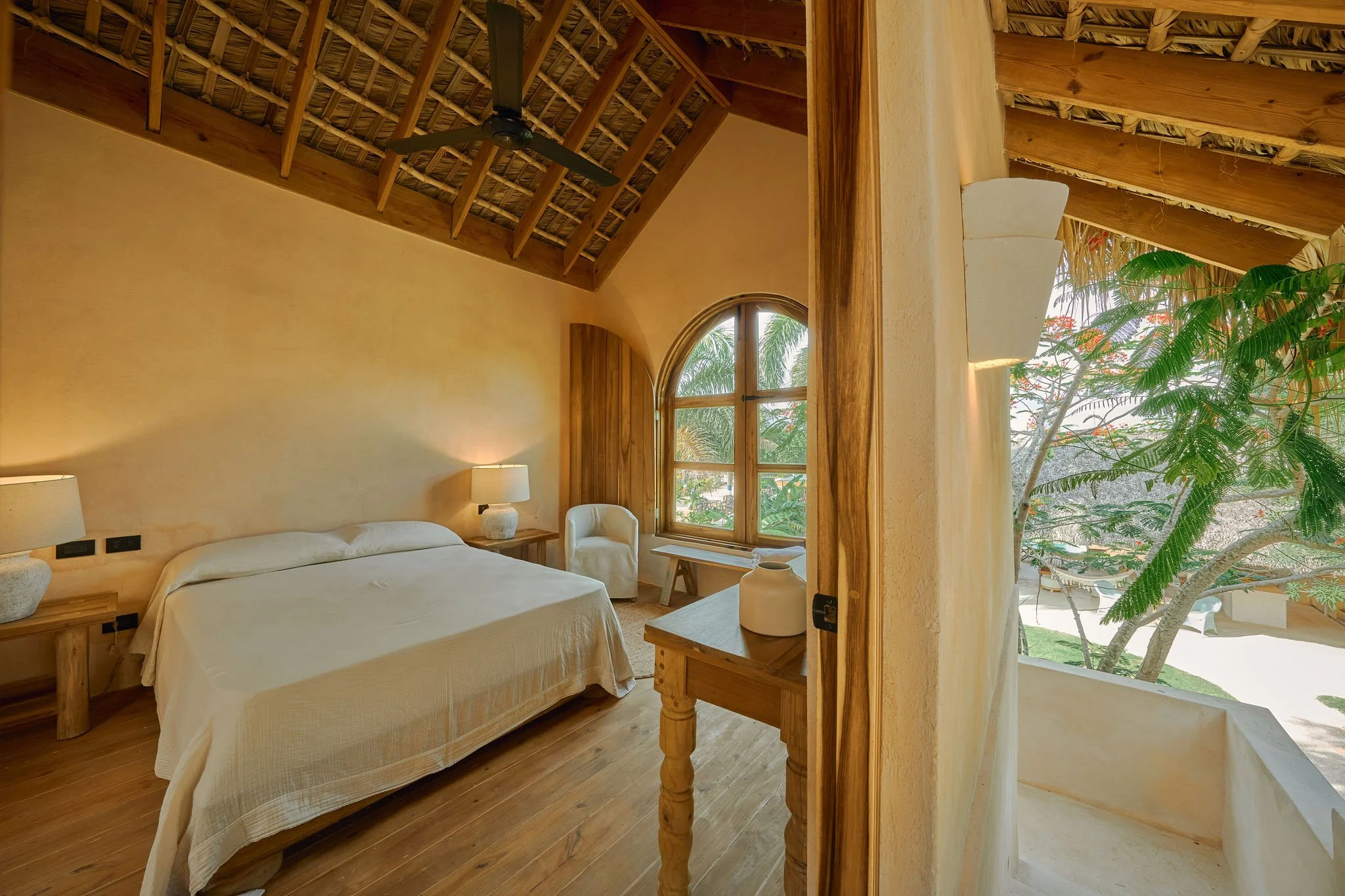 Habitación decorada con muebles de madera, cama con sábanas blancas, lámparas en las mesitas, ventana con vista a vegetación y balcón con plantas tropicales.