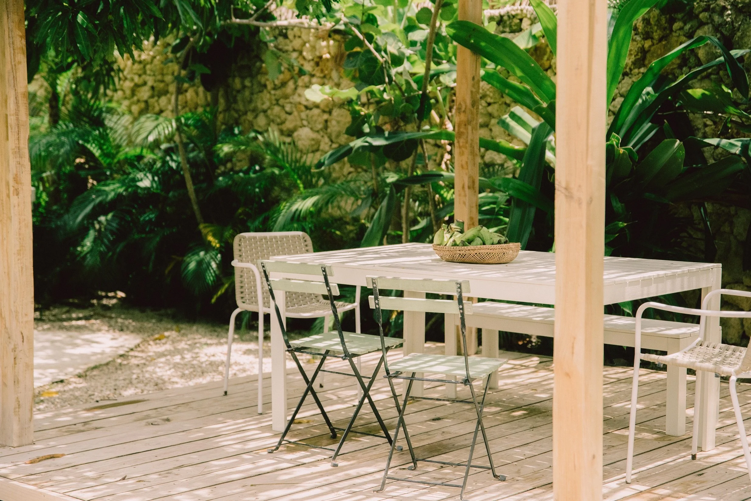 Mesa de madera blanca con sillas blancas y negras en un espacio abierto con plantas verdes y una pared de piedra en el fondo.
