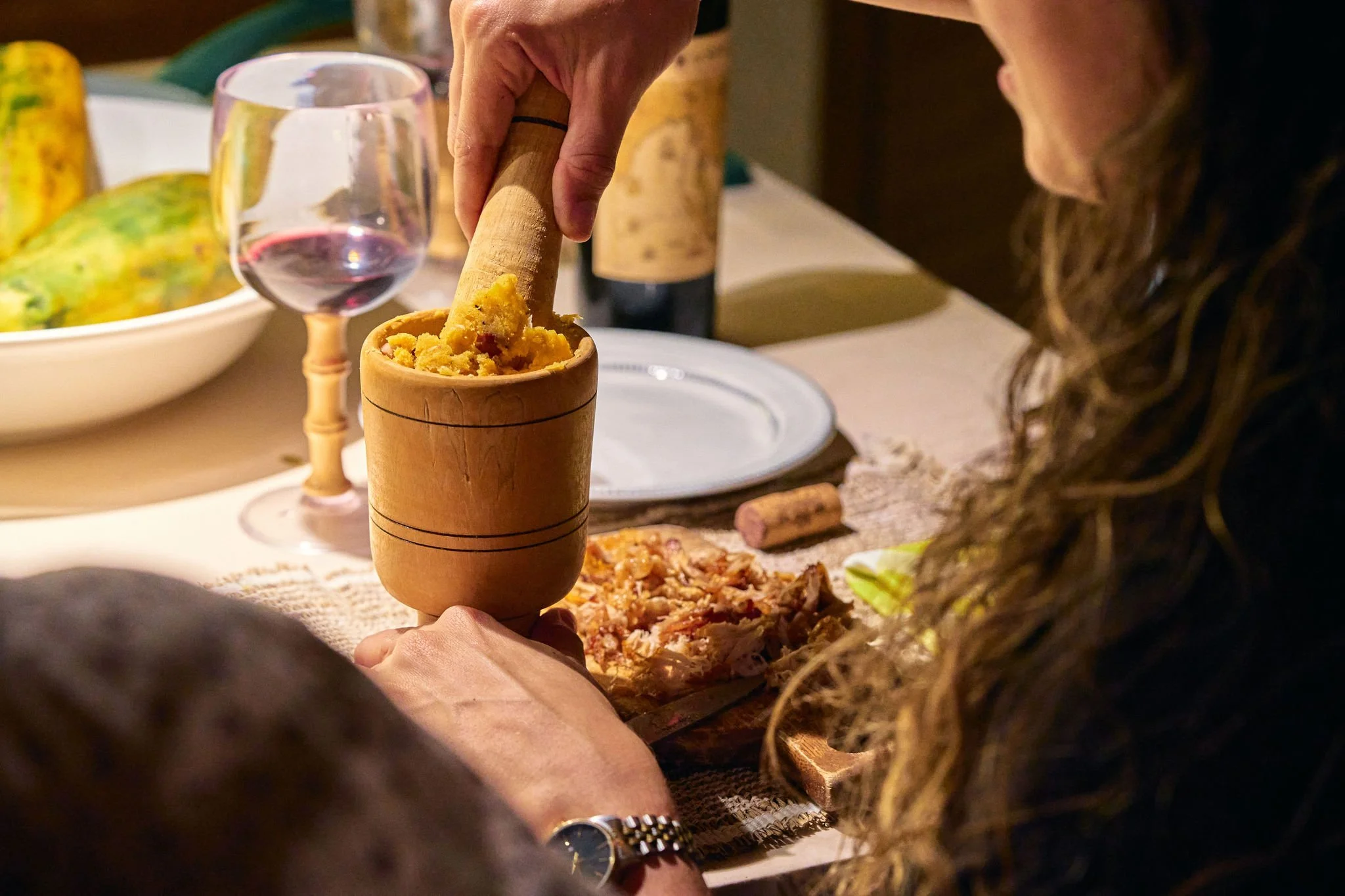 Persona machuca con un mortero y maja una masa amarilla en una mesa rodeada de comida y bebidas, incluyendo vino y una fruta grande y amarilla.