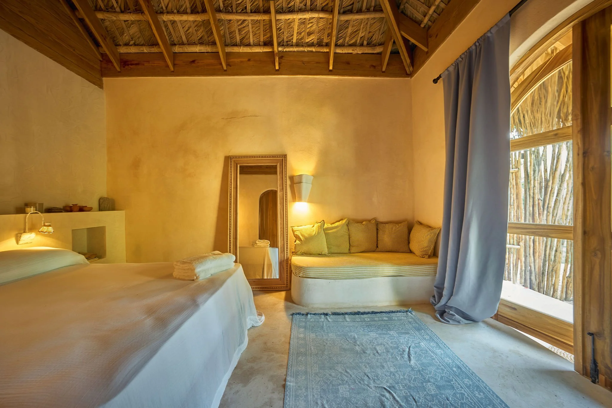 Habitación con cama, espejo de marco dorado, sillón con cojines, cortinas y ventana de madera con vista a palmeras.