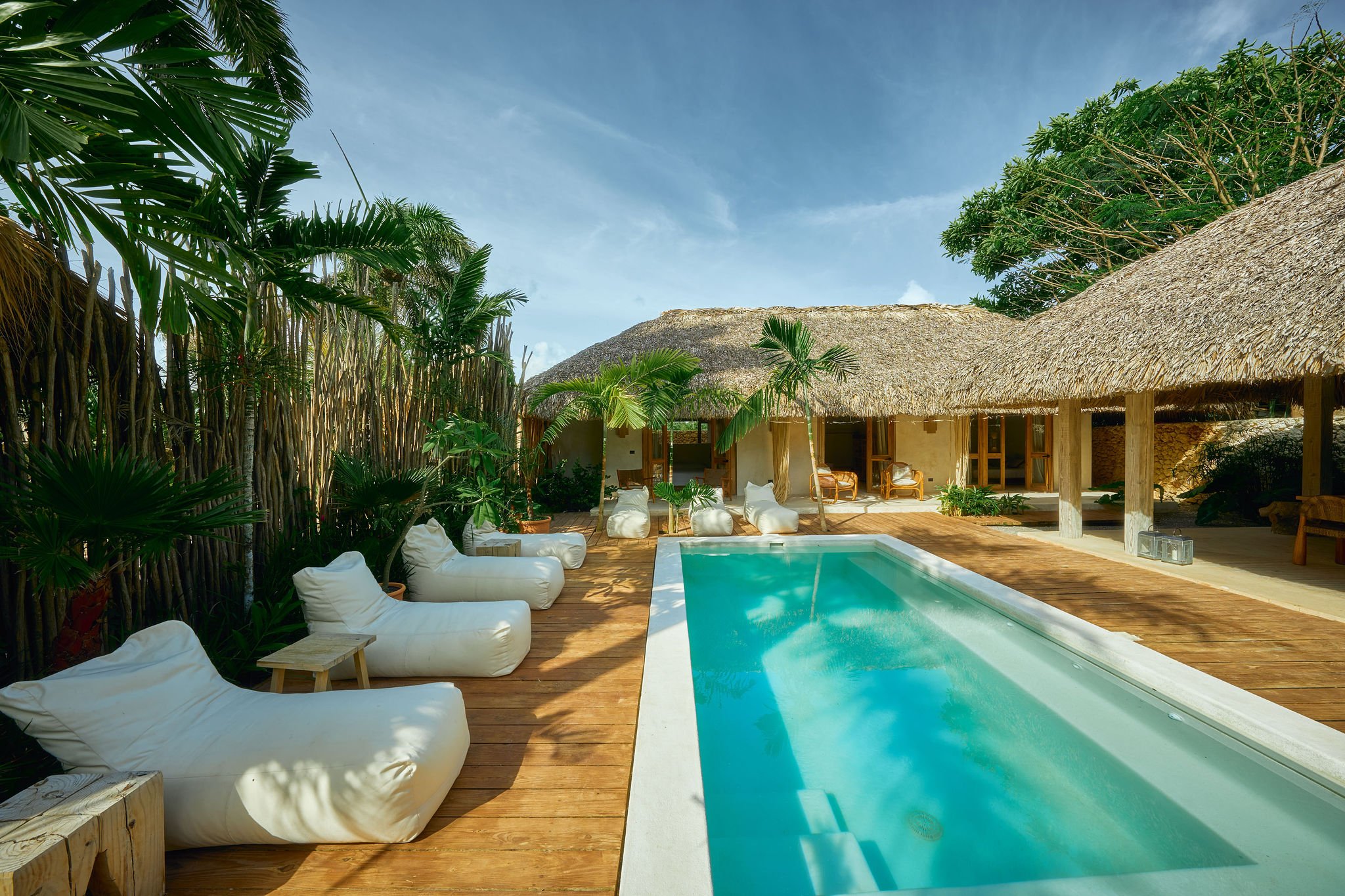 Piscina en una cabaña de playa rodeada de plantas tropicales y muebles blancos.
