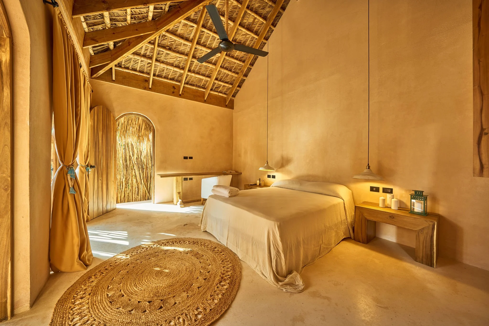 Habitación con paredes de color beige, un techo de madera y un ventilador. Hay una cama con sábanas blancas, una lámpara colgante a cada lado, una mesita con velas y un objeto decorativo. En la esquina izquierda, cortinas doradas y una ventana con pe