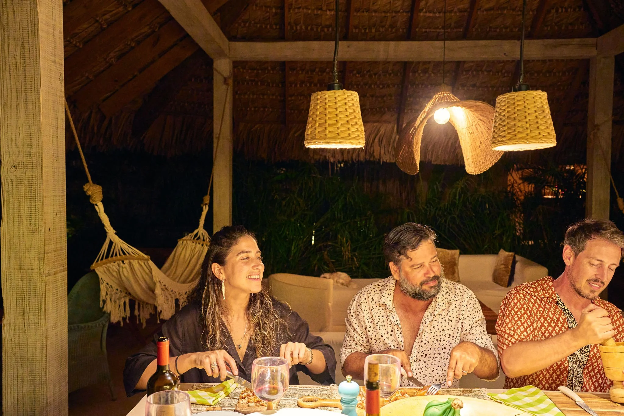 grupo de personas cenando en un ambiente acogedor con decoración rústica, lámparas de madera, hamaca y plantas en el fondo, disfrutando de una comida.