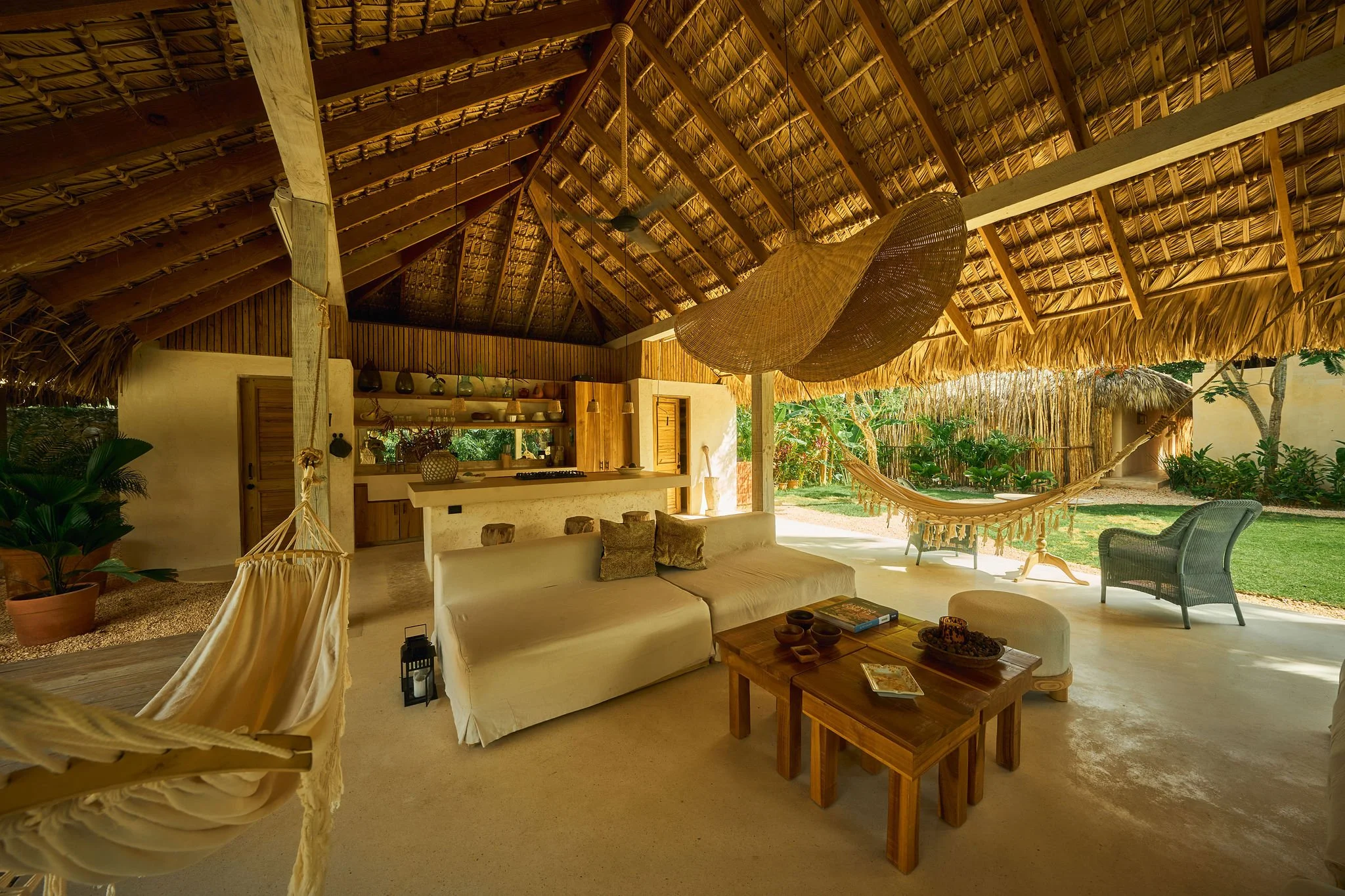 Interior de una sala con techo de palma, sillón, hamaca, mesas de madera, y vista al jardín exterior con césped y plantas