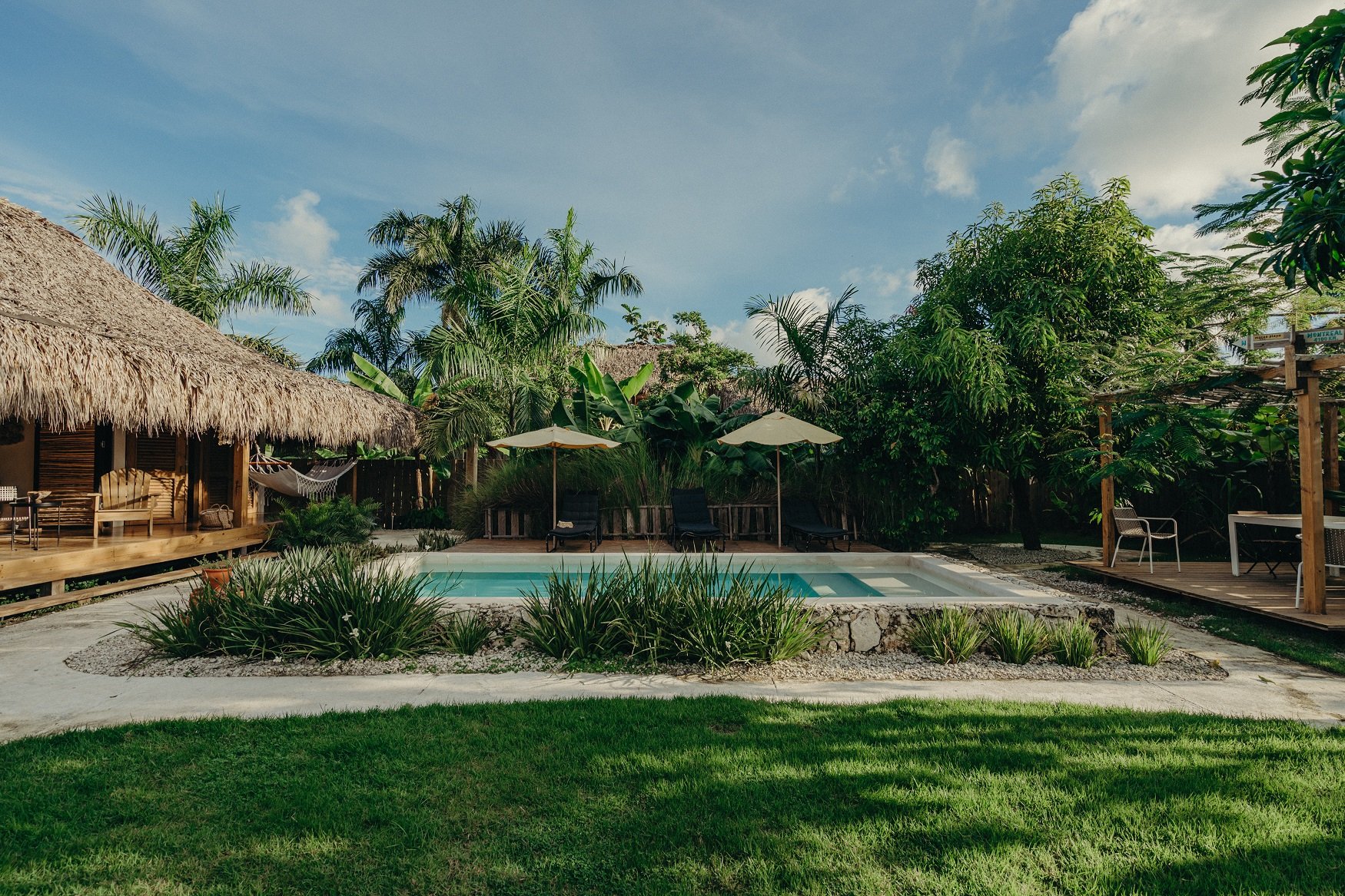 Patio con piscina pequeña, sombrillas, sillas, hamaca, y estructura de madera rodeada de plantas tropicales en un día soleado con cielo azul
