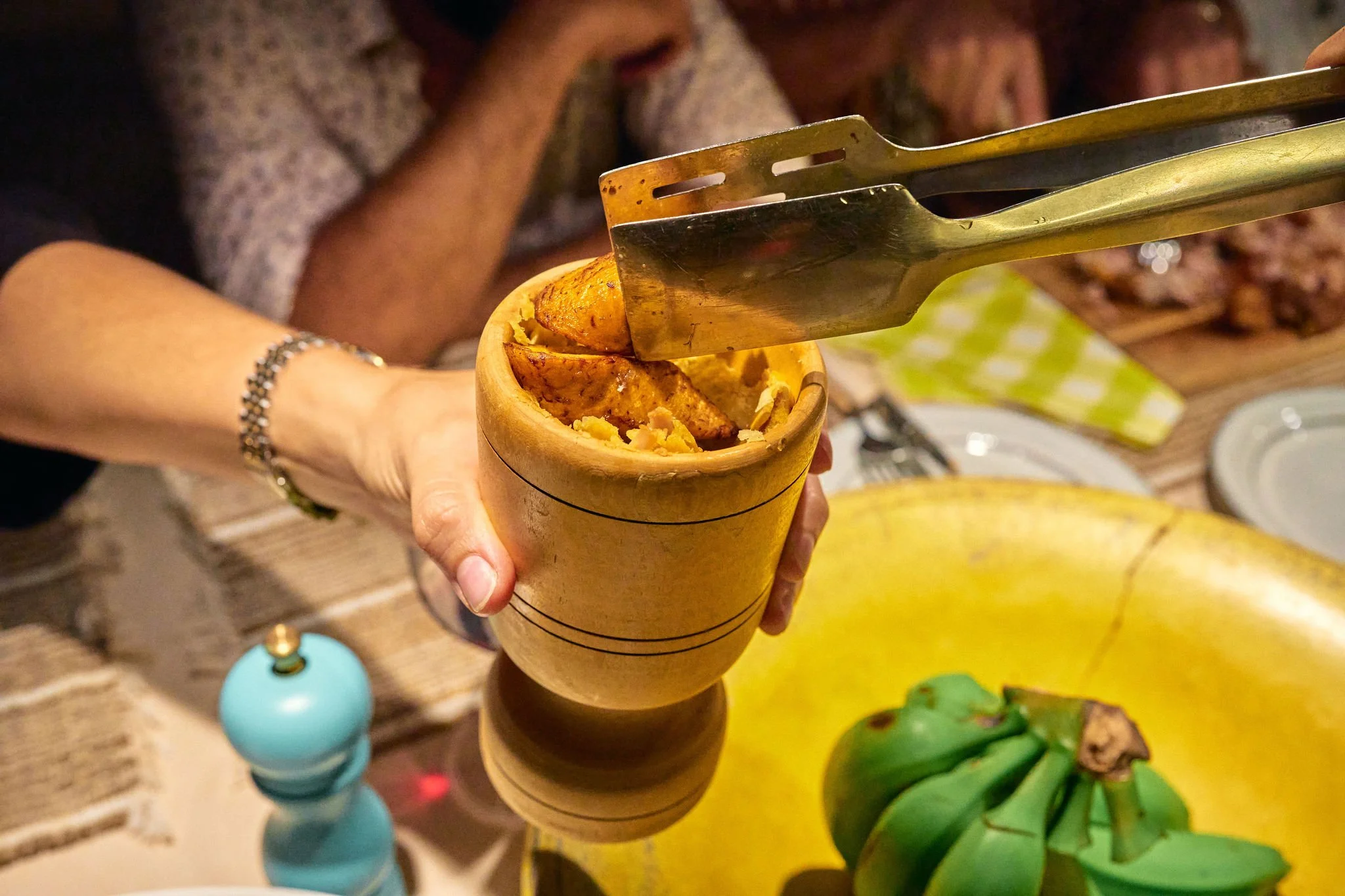 Una persona con una pulsera en la muñeca sostiene un recipiente de madera con comida y está usando pinzas para colocar un plato de comida en un recipiente redondo en una mesa.