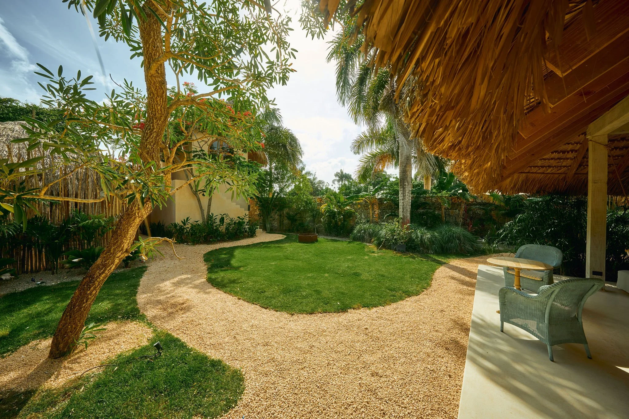 Jardín con sendero de grava, césped y árboles tropicales, con una terraza de madera con sillas y mesa bajo un techo de hojas secas.
