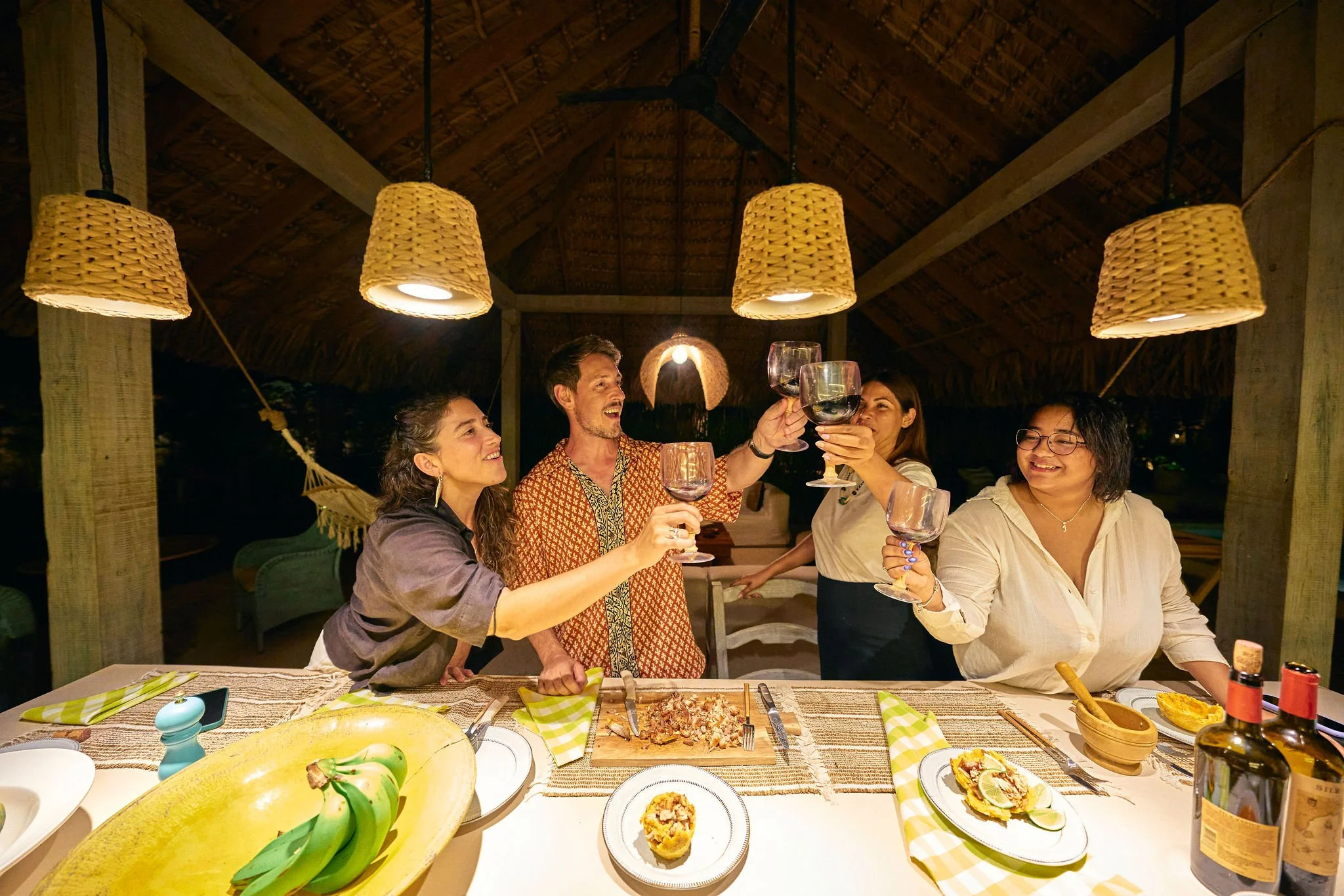 Grupo de personas brindando en una cena al aire libre en una estructura de madera con techo de paja, con comida y vino en la mesa.
