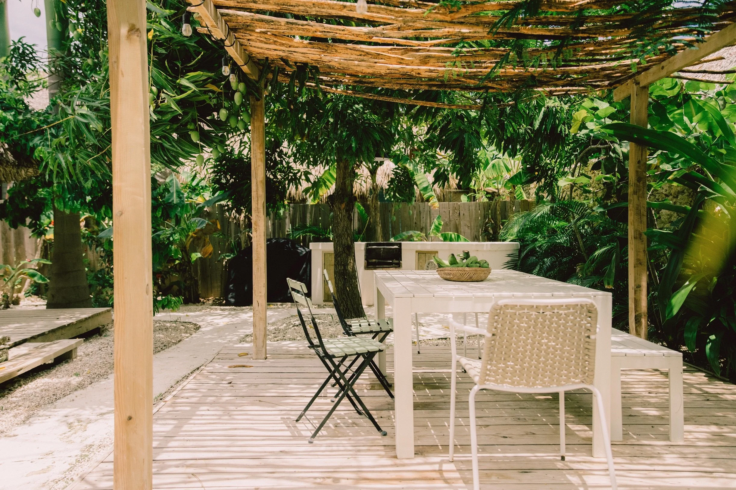 Un área de comedor al aire libre con mesa blanca y sillas en un entorno rodeado de vegetación tropical y sombra parcial con estructura de madera y cobertura de ramas.