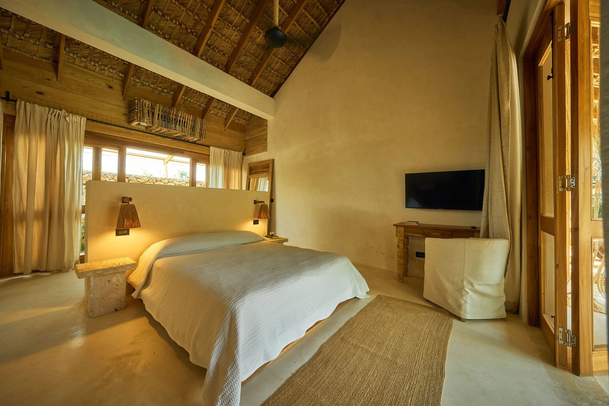 Habitación con paredes de color crema, cama con sábanas blancas, dos lámparas de pared, cortinas blancas, ventanas grandes con vista al exterior, un mueble para televisión, una silla y una puerta de vidrio que da a una terraza con vista natural.