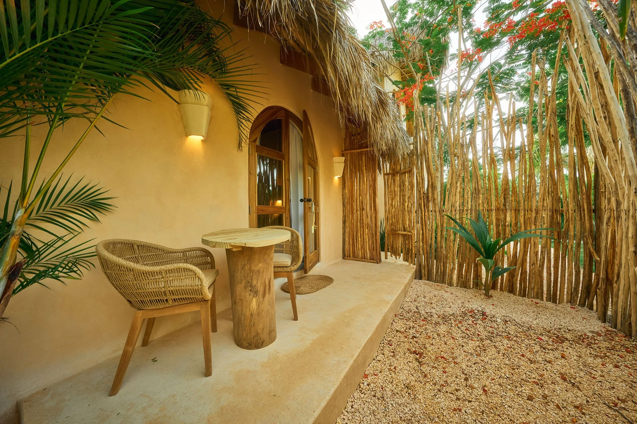 Un patio exterior con paredes de color crema, una puerta de madera con arco, sillones y mesa de madera, y una cerca de cañas de bambú que rodea el espacio, con plantas tropicales y flores.