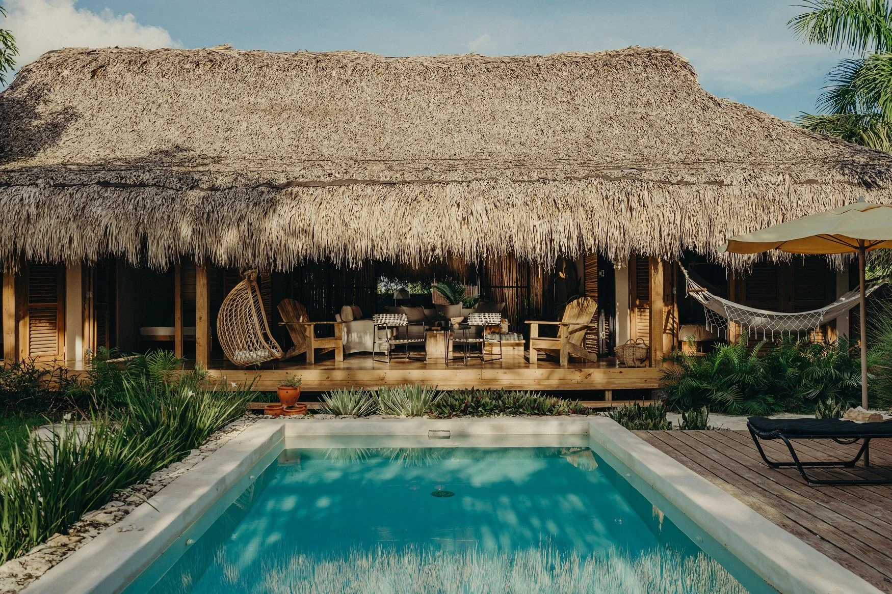 Casa de playa con techo de paja, piscina y área de descanso con sillas y hamaca, rodeada de plantas tropicales.