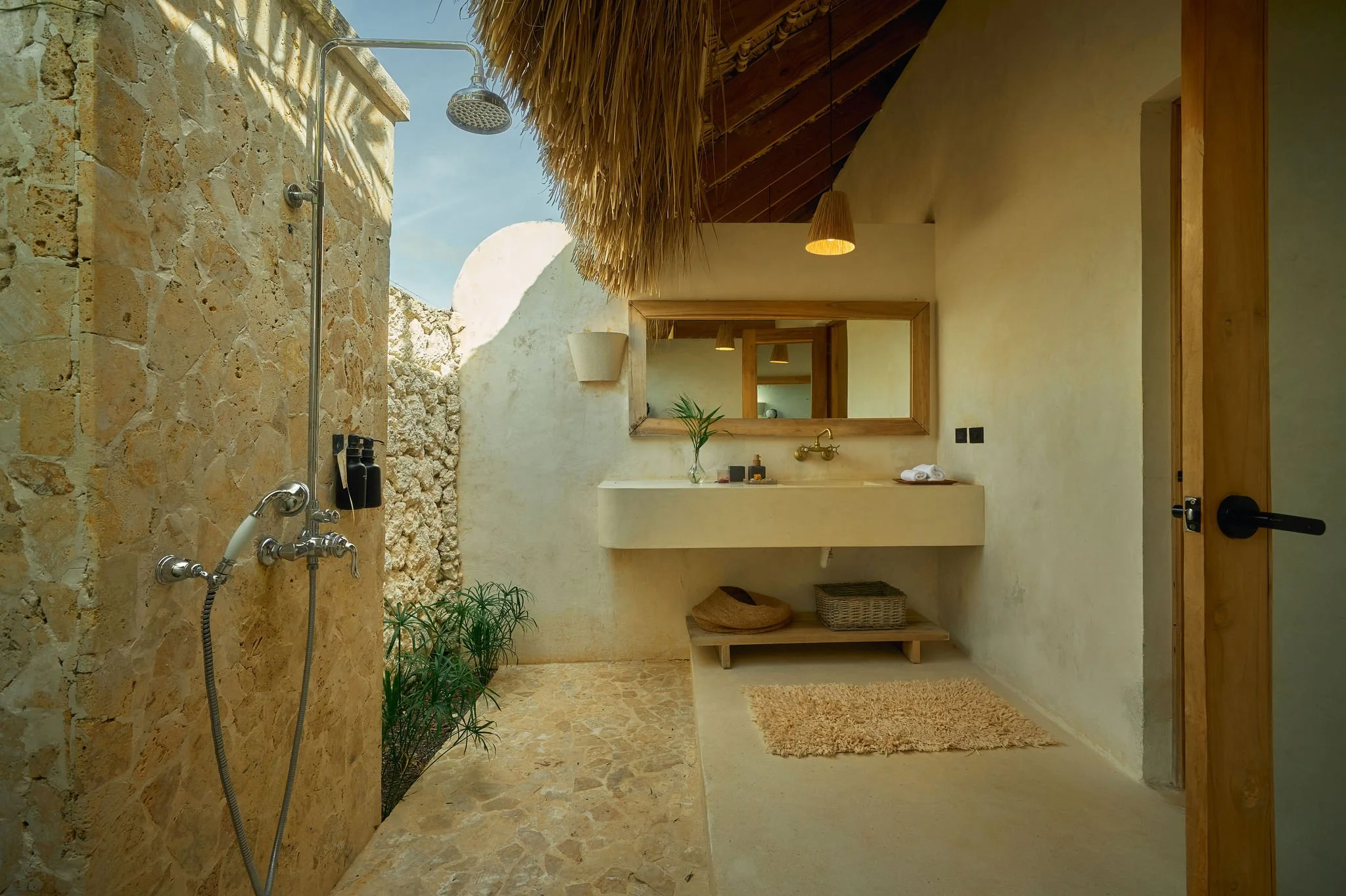 Baño con ducha al aire libre, paredes de piedra y techo de paja, espejo grande, lavabo con accesorios, pequeñas plantas y alfombra de baño